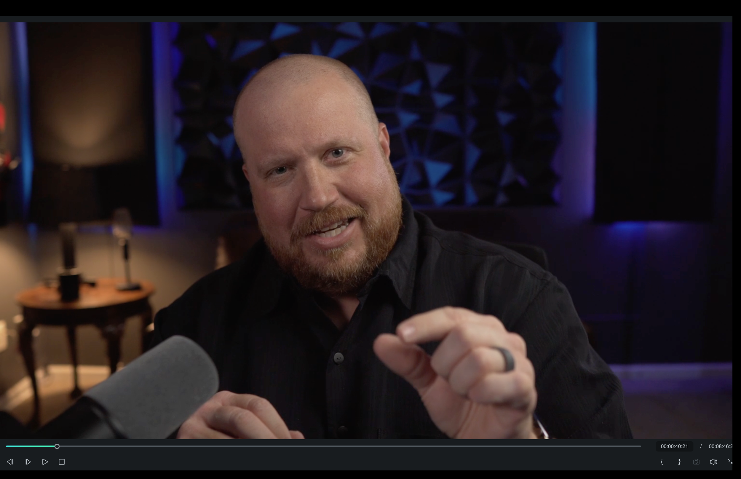 A man with a beard and mustache speaking into a microphone in a dimly lit room with blue and purple lighting, seated at a table.