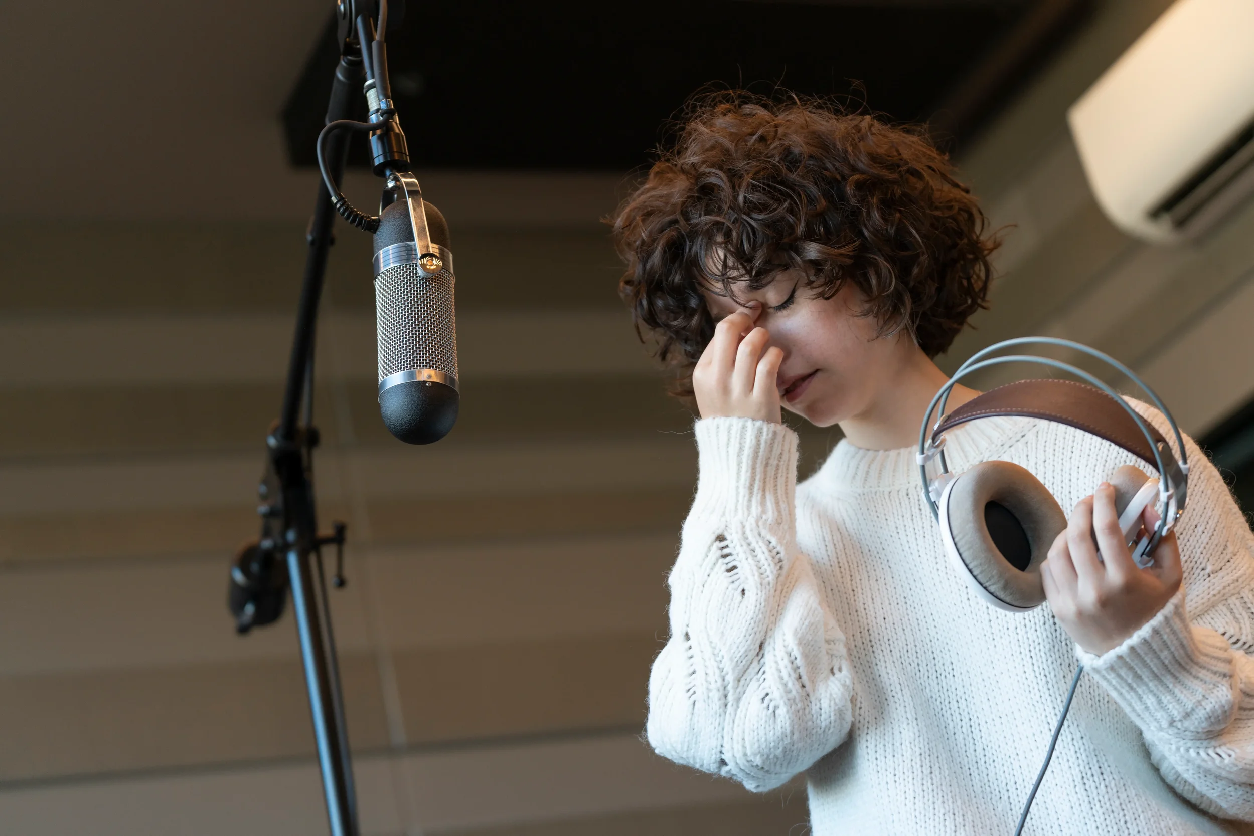 A woman with curly hair in a white sweater holding headphones and pinching her nose while standing in front of a microphone in a recording studio.