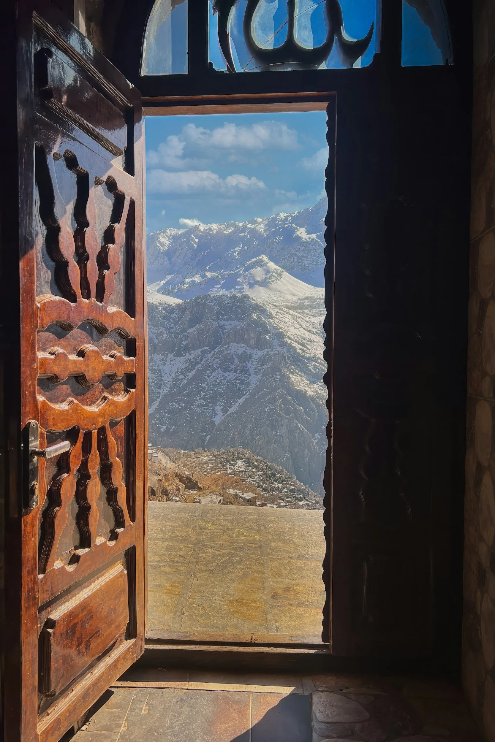 Open wooden door leading to a mountain landscape with snow-capped peaks and a clear blue sky.