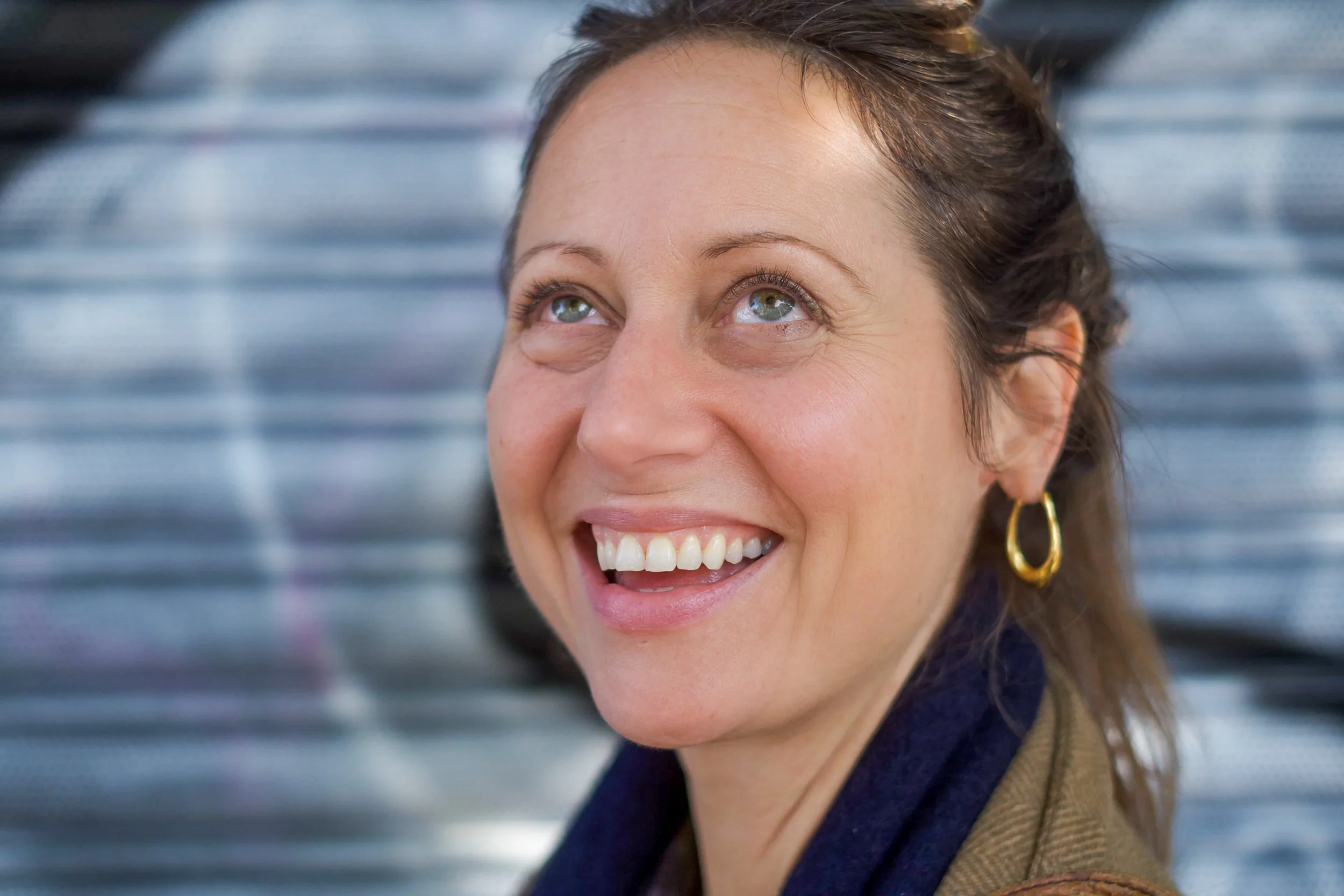 Close-up of a woman with light skin and brown hair smiling and looking up, wearing gold hoop earrings and a brown jacket, with a blurred metallic background.
