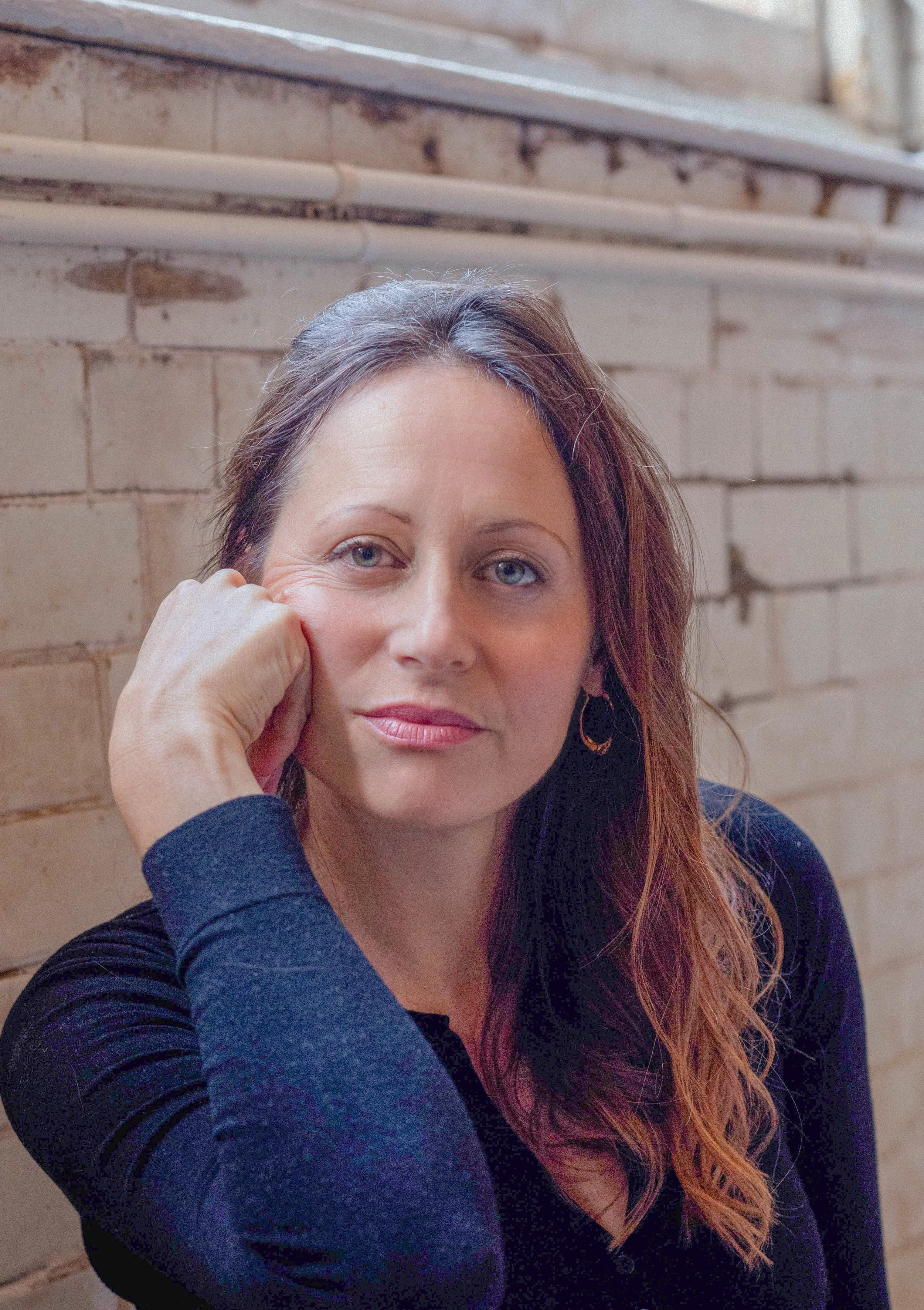 A woman with long, wavy chestnut hair and green eyes sitting against a brick wall, resting her face on her hand, wearing a black long-sleeve top and hoop earrings.
