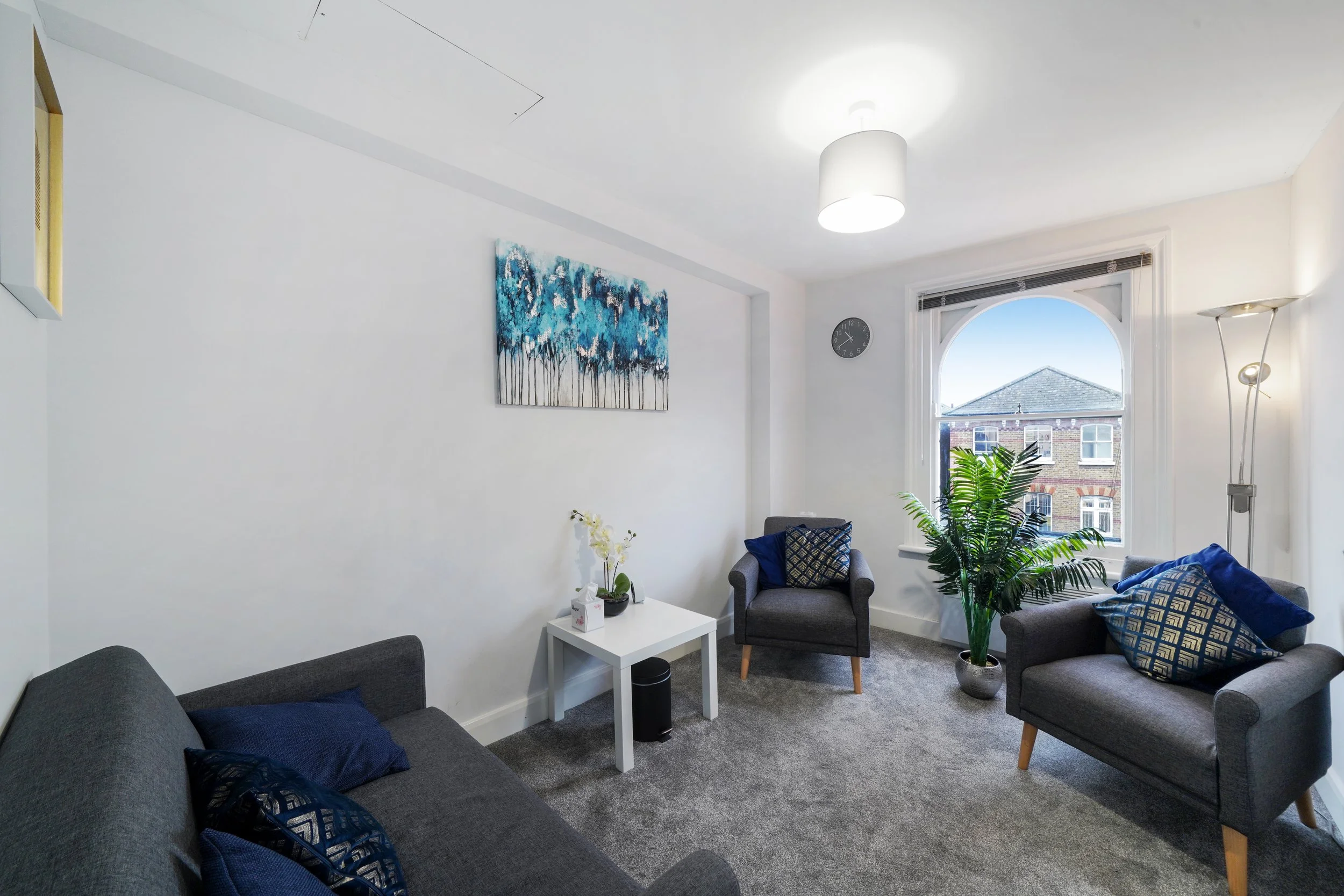 A therapy clinic room with three grey armchairs, a small white table, a large window, a green potted plant, and wall art featuring blue trees.