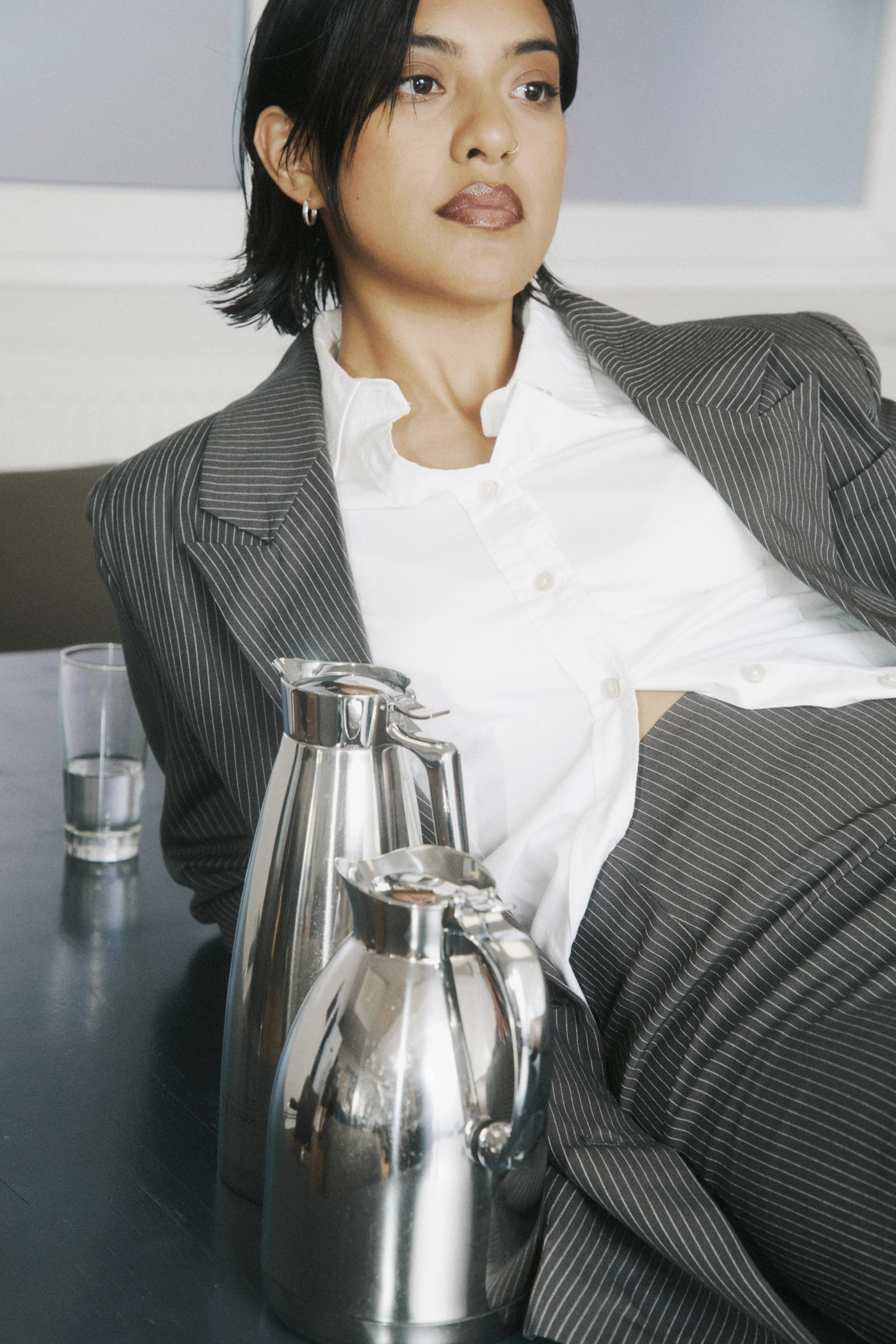 A woman with short black hair and earrings, dressed in a suit with a white shirt, sitting at a table with a glass of water and metallic thermoses.