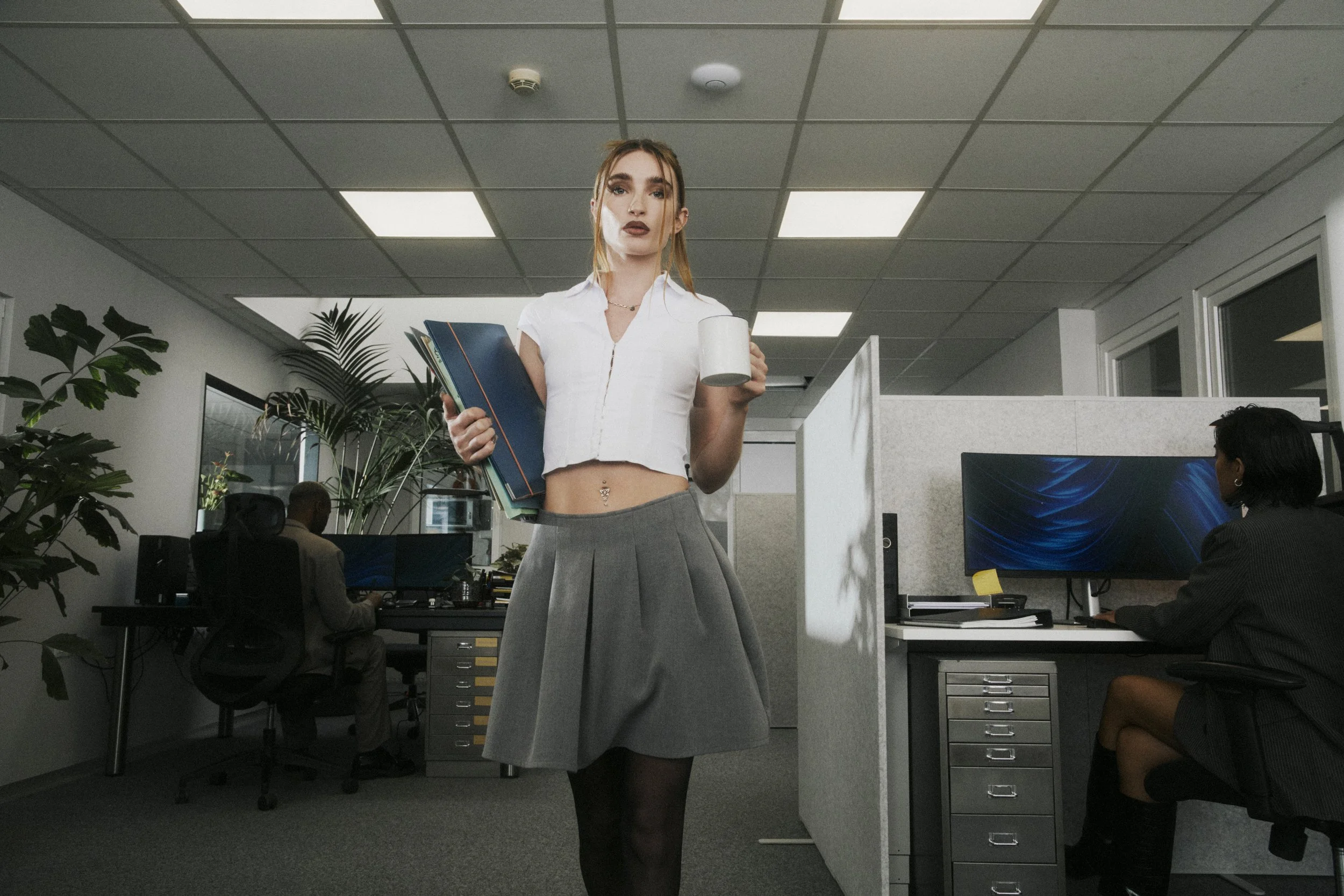 A woman in a white cropped blouse and gray pleated skirt walking through an office while holding a mug in one hand and a stack of folders in the other, with two other people working at desks in the background.