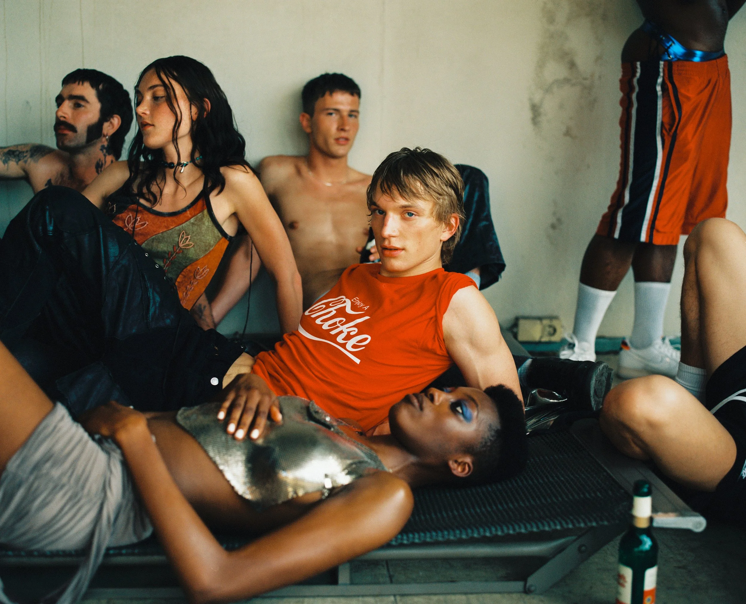 Group of young people relaxing in a room, some lying on a cot and others sitting against the wall, with casual attire including vintage and sporty clothing.
