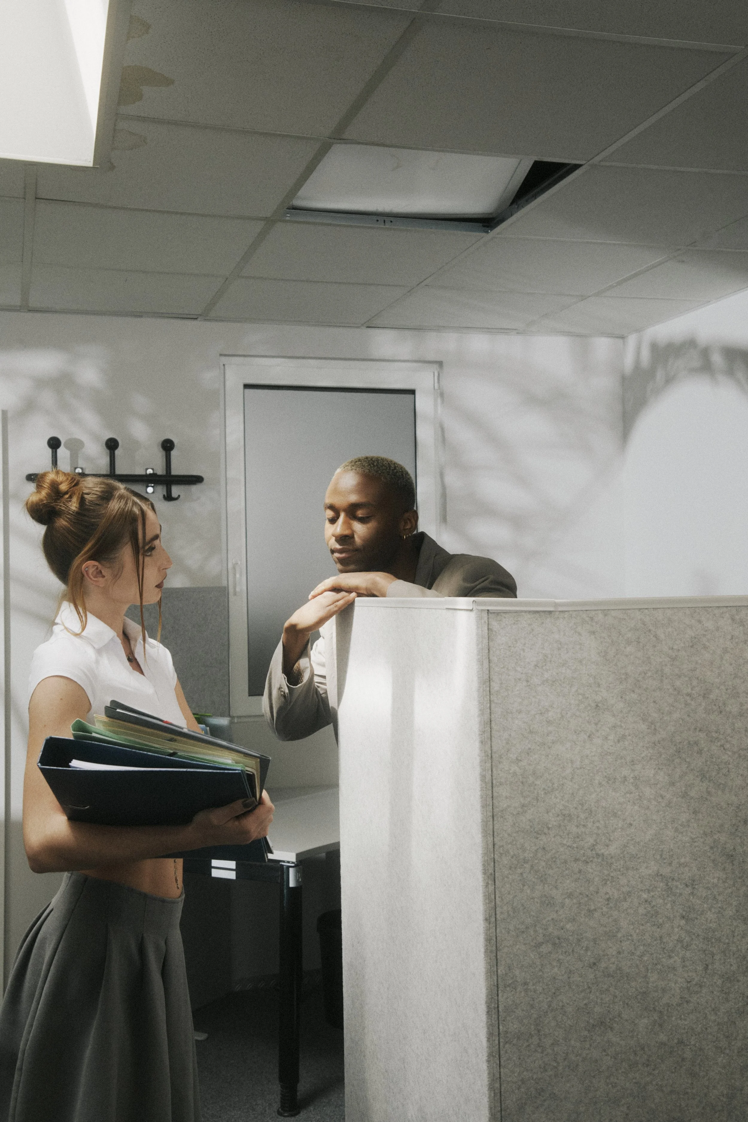 A woman in office attire holding a folder with several documents in an office cubicle, talking to a man leaning on the cubicle wall.