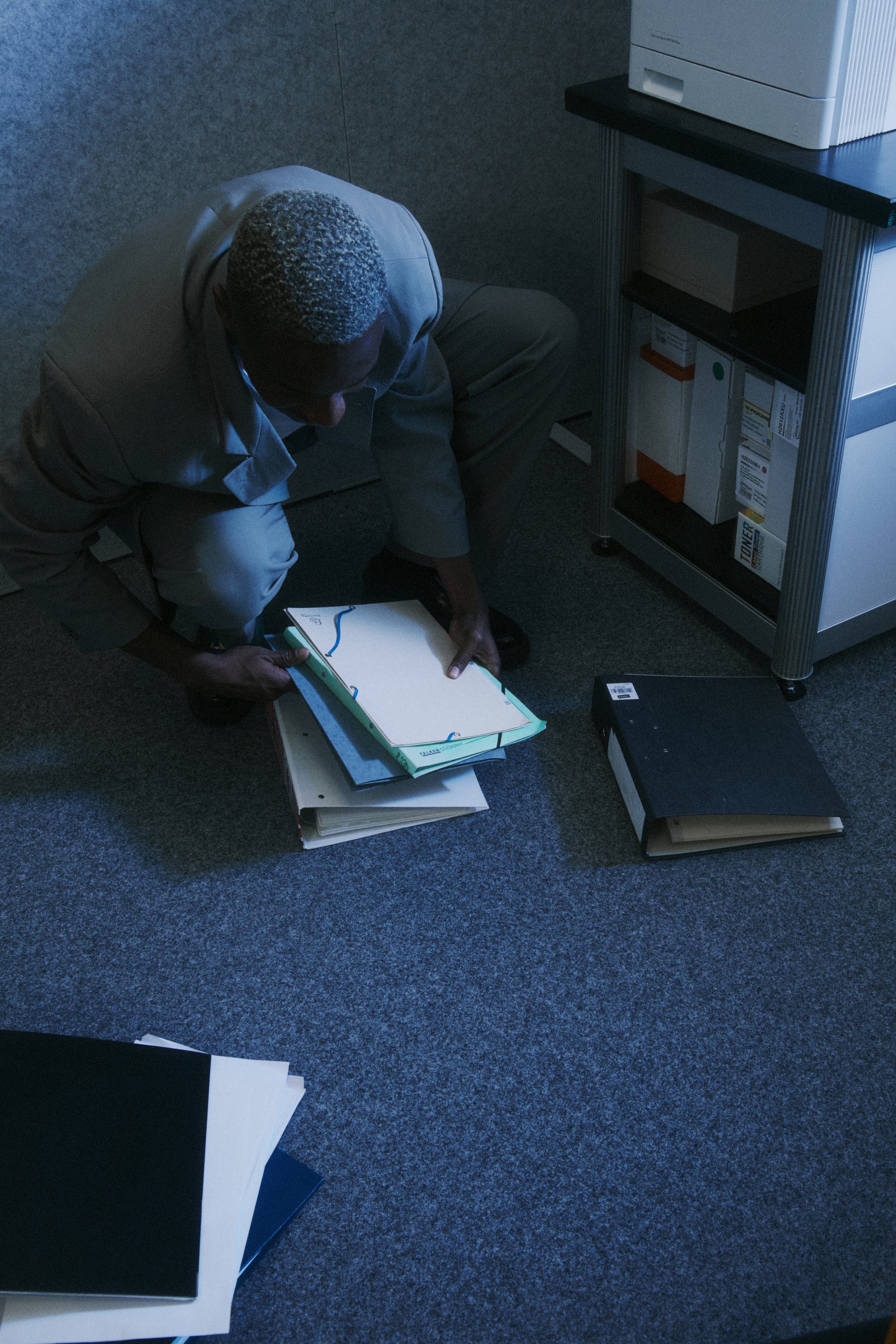 A man with short, salt-and-pepper hair wearing a gray suit is kneeling on a gray carpeted floor, organizing various binders and folders in an office. There is a small shelf with office supplies and a printer beside him.