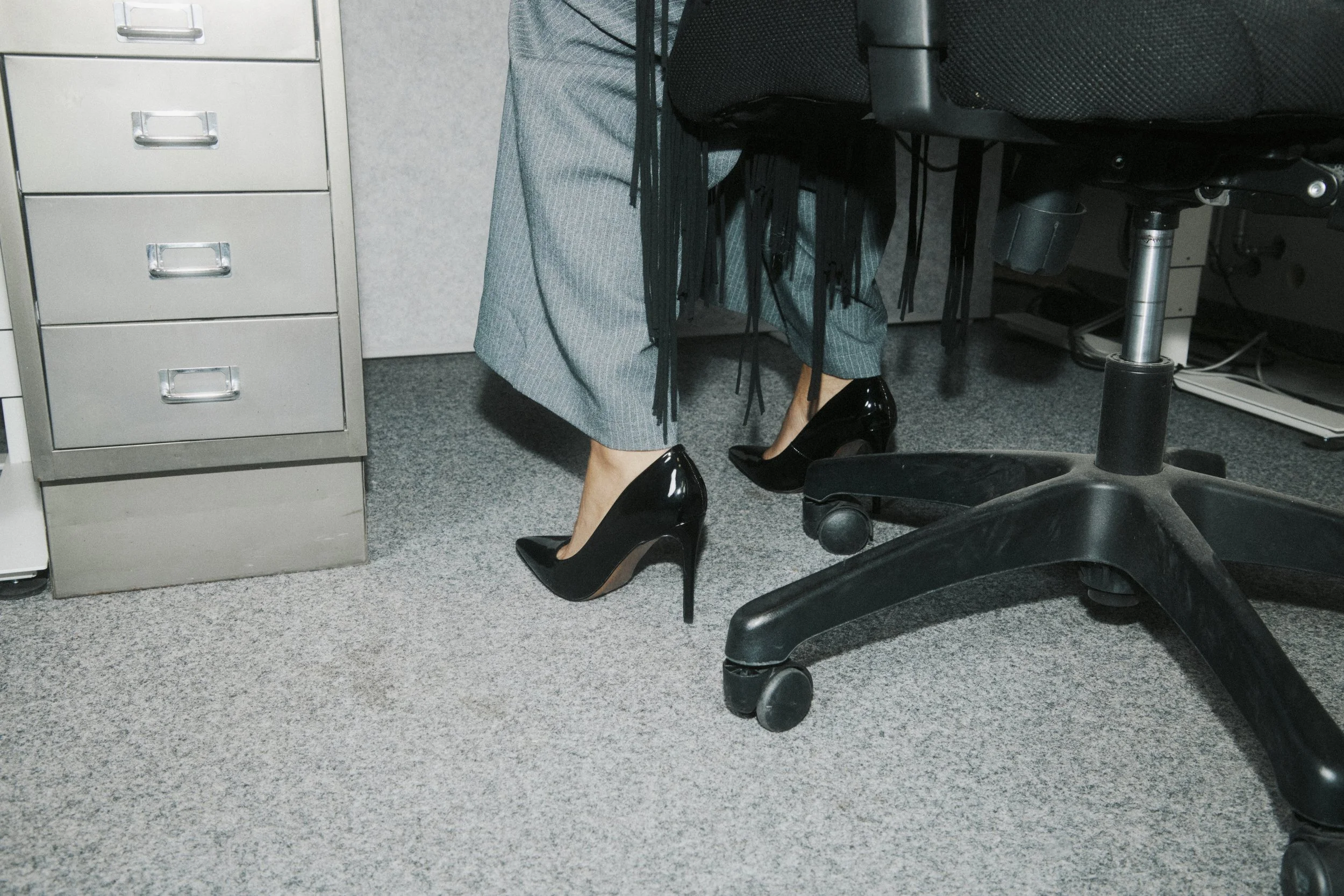 Office setting showing a person sitting at a desk wearing checked pants, black high heels, and a fringed black bag. A gray filing cabinet is on the left, and a black office chair with wheels is on the right.