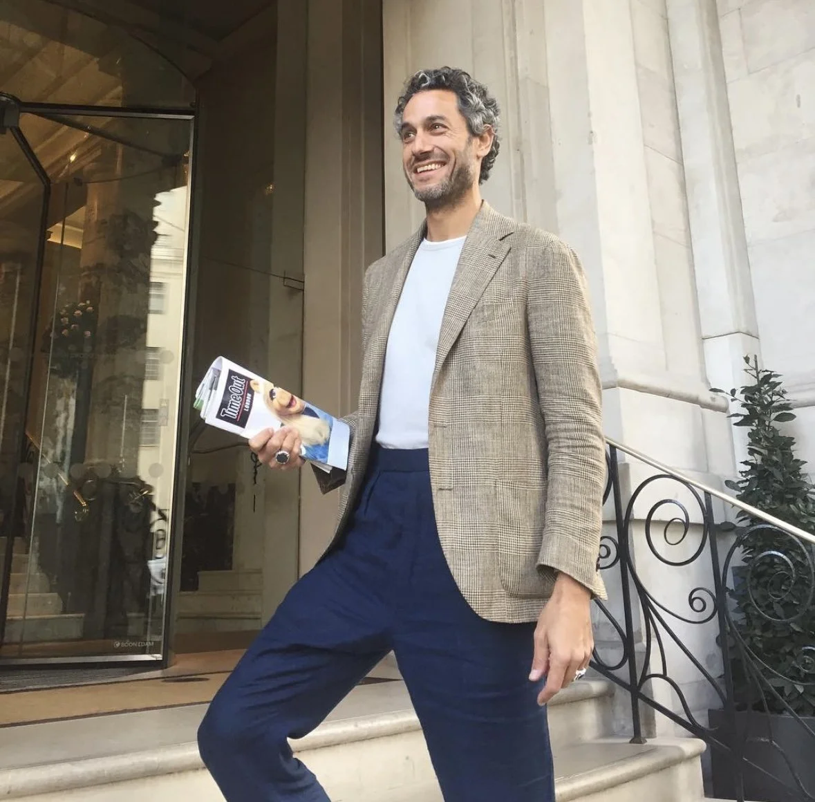 A man with gray curly hair, beard, wearing a beige plaid blazer, white t-shirt, and navy blue pants, standing outside a building holding a magazine.