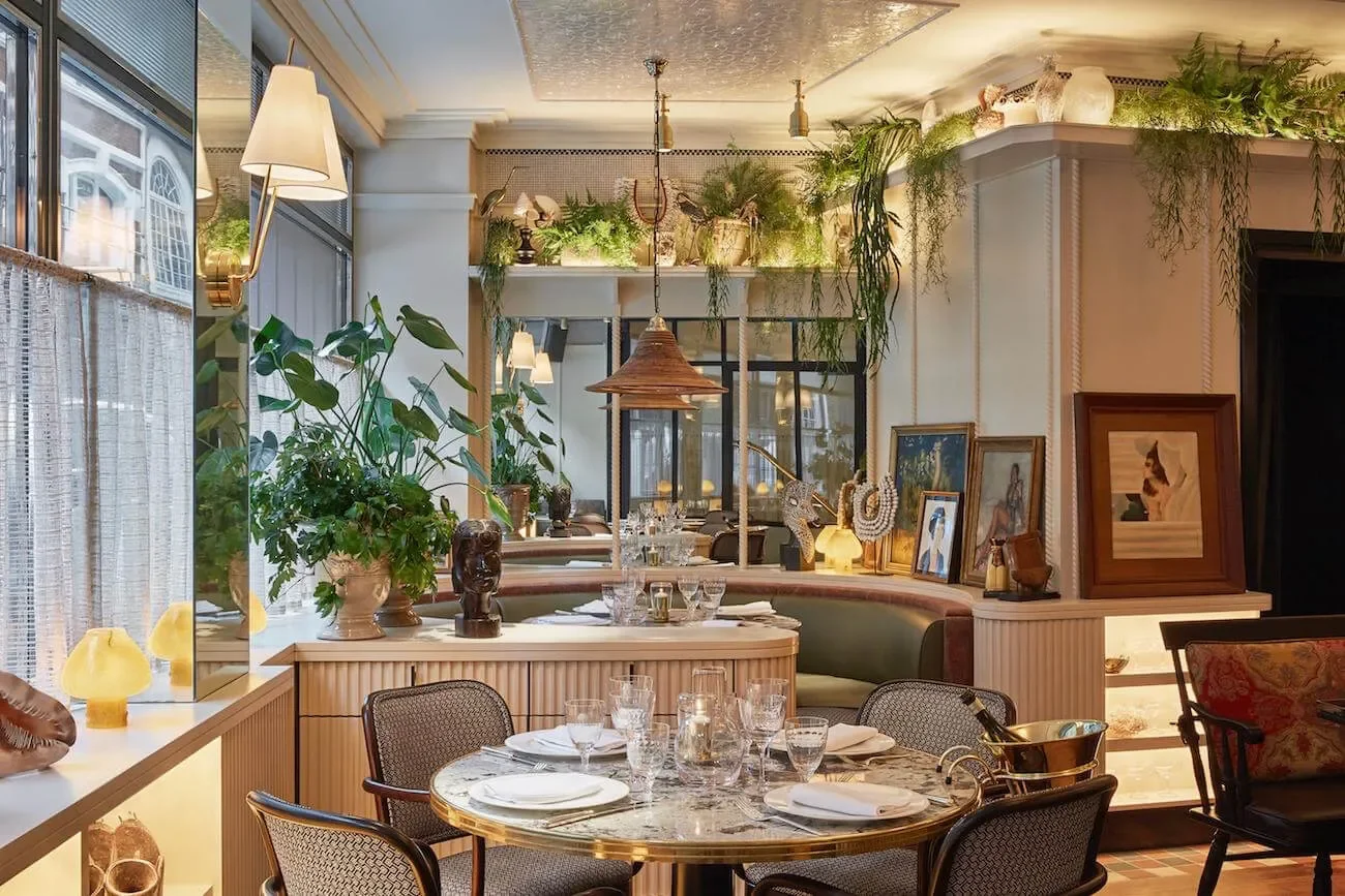 Indoor restaurant dining area with a round marble table set for a meal, surrounded by upholstered chairs. The space features large windows, green plants, framed artwork on the walls, and decorative lighting fixtures.