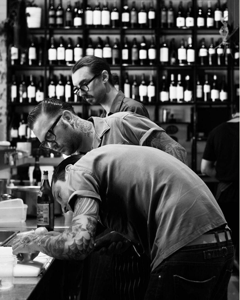 Three tattooed men working together in a kitchen or restaurant setting, with shelves of wine bottles in the background.