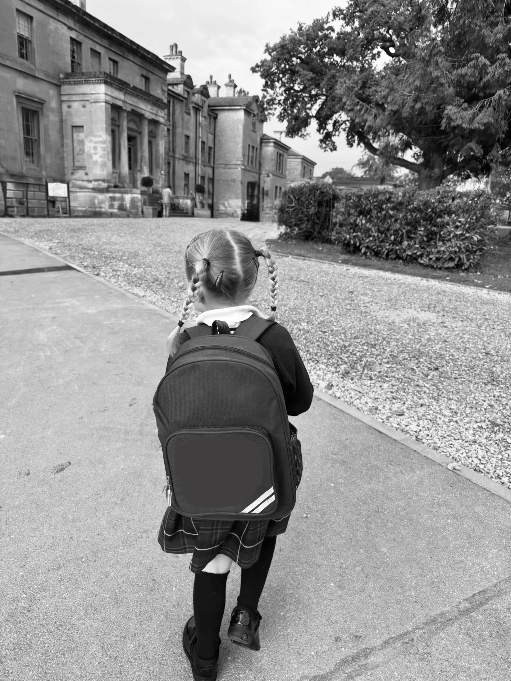 Child with pigtails and a backpack walking on a school sidewalk near historic buildings and trees.