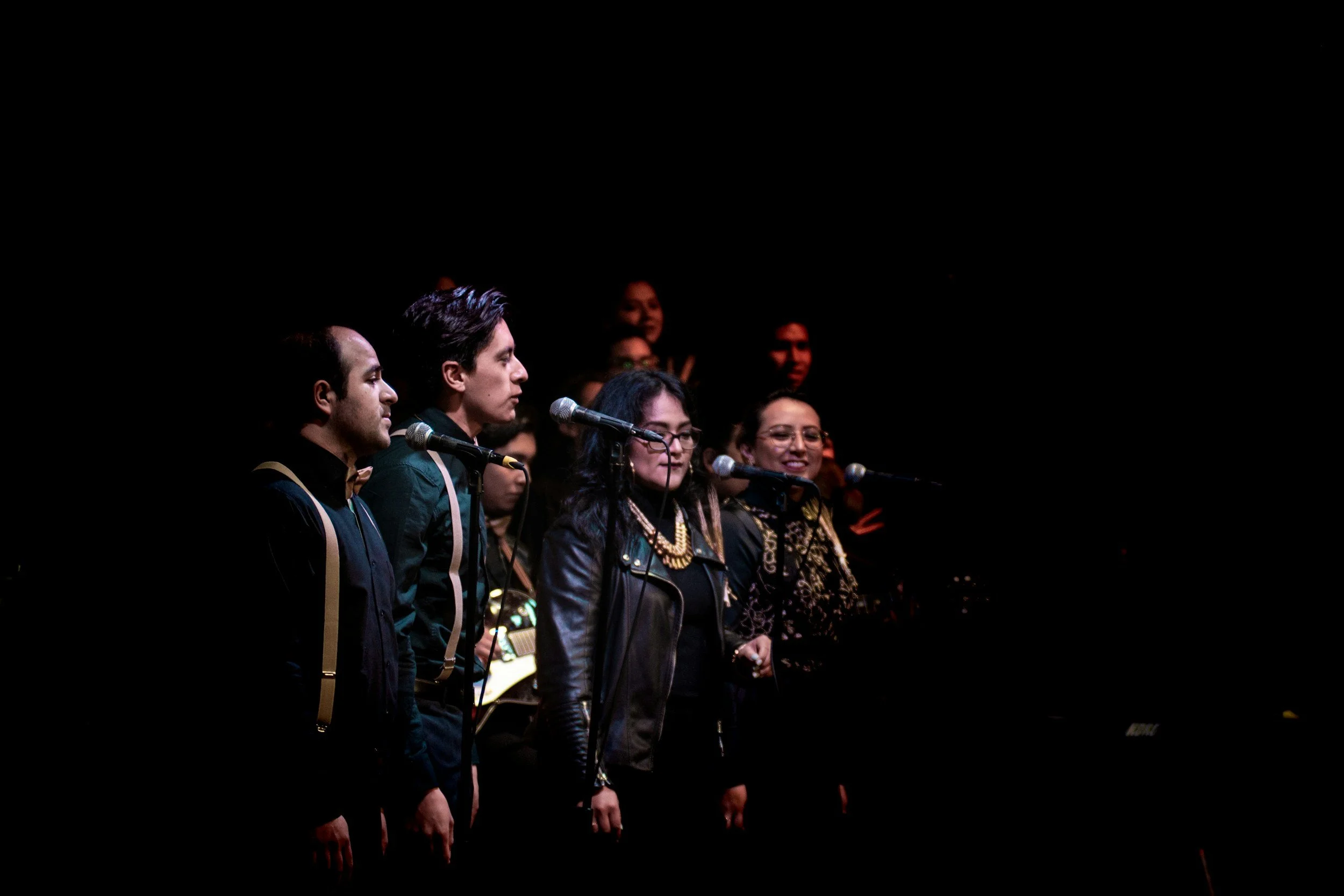 A group of performers standing in a line on stage, with some holding microphones, illuminated by stage lighting, against a dark background.