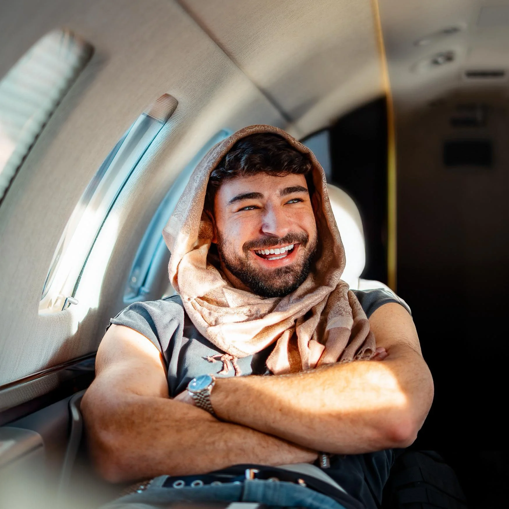 A smiling man with a beard and dark hair, sitting inside an airplane with arms crossed, wearing a hooded scarf and a watch.