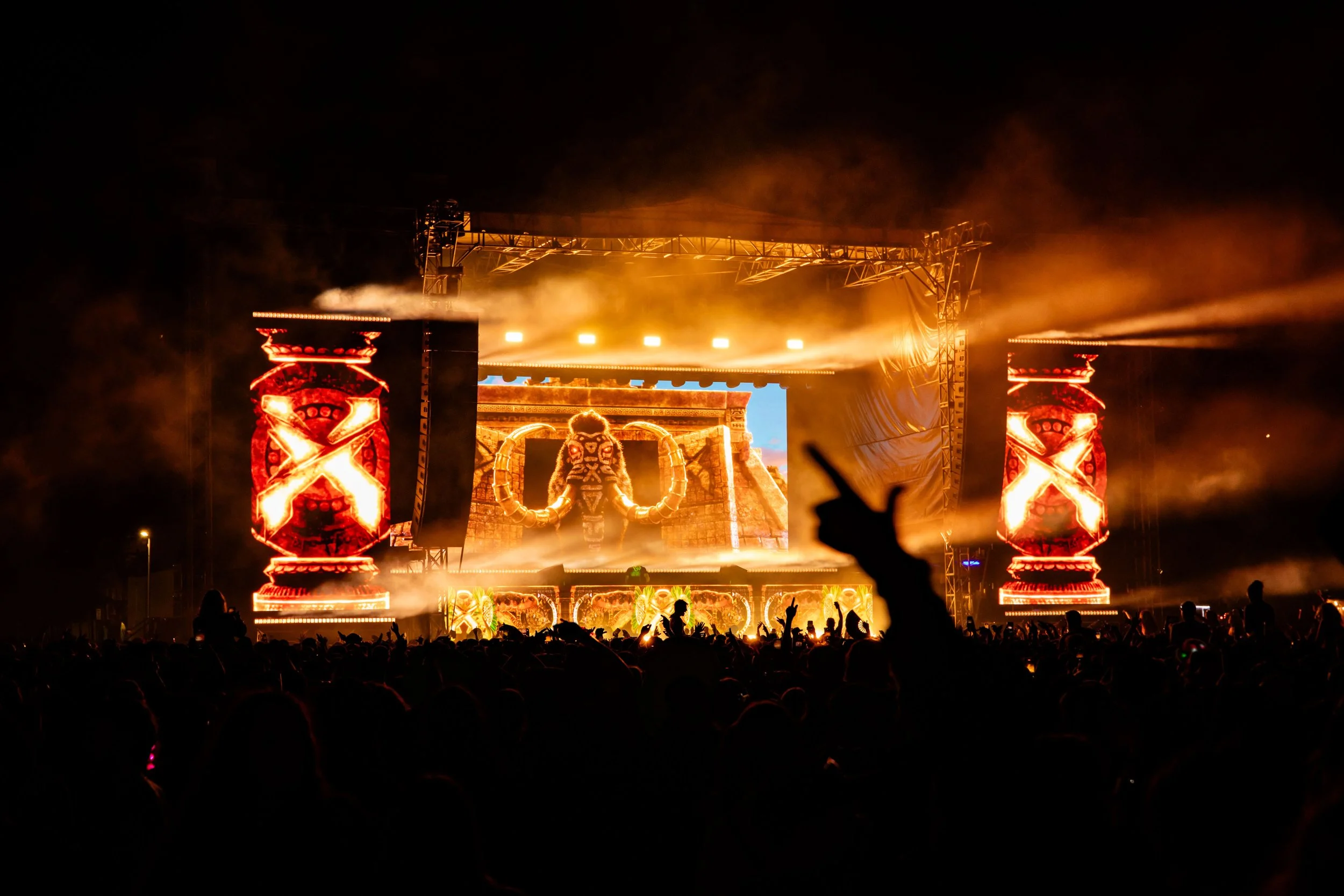 A large outdoor concert stage with bright orange and yellow lighting, digital screens displaying tribal imagery, and an audience with hands raised at night.