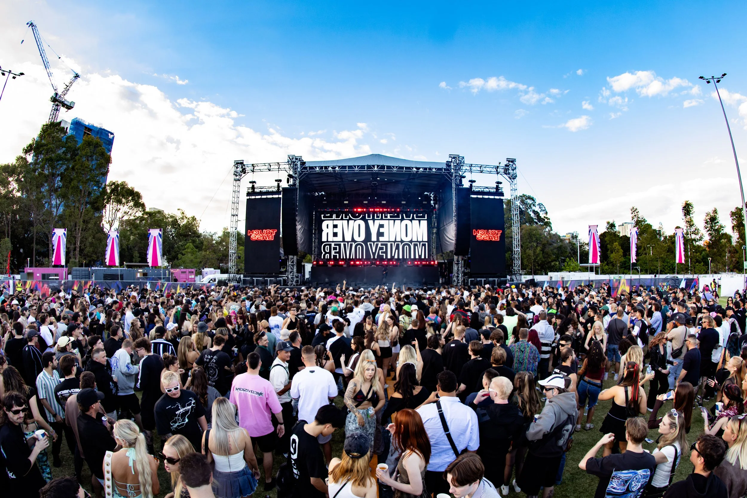 Crowd of people at an outdoor concert or festival with a large stage, screens, and trees in the background on a clear day.