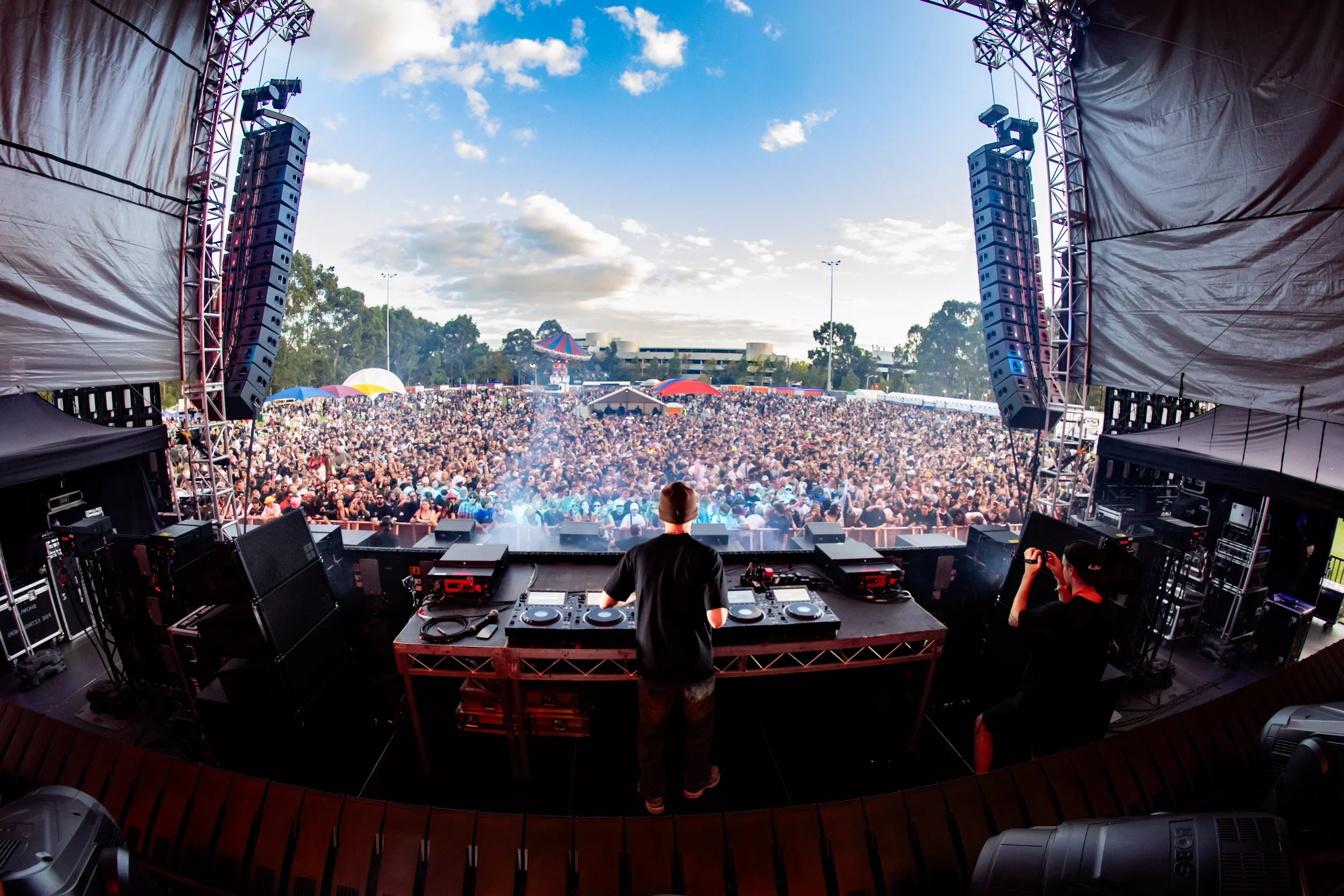 A DJ performs on stage at an outdoor music festival with a large crowd in the background under a partly cloudy sky.
