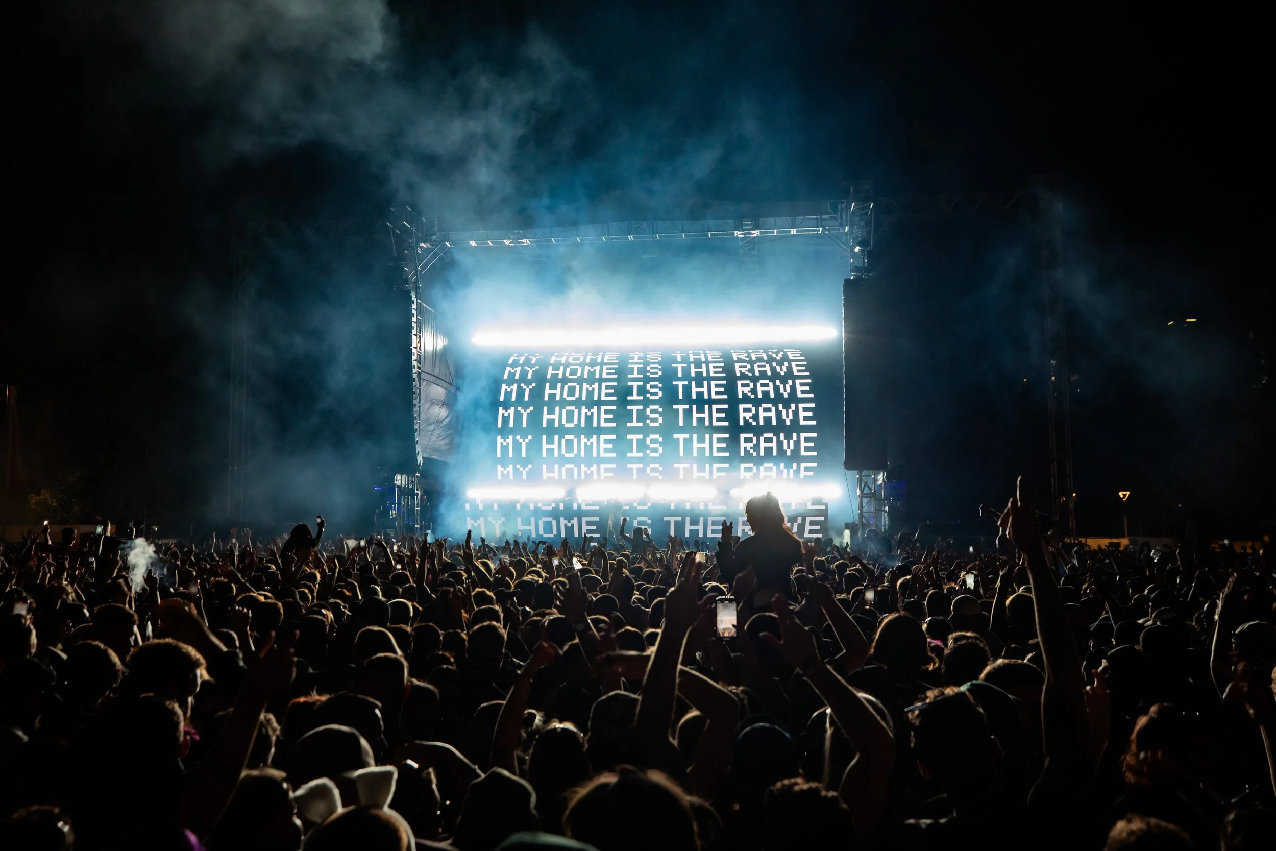 Crowd at a nighttime concert or music festival with a large illuminated screen displaying lyrics: 'MY HOME IS THE RAVE.'