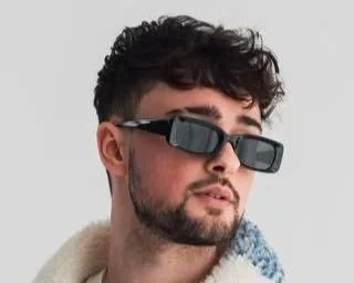 A man with dark hair, sunglasses, and a beard wearing a light-colored shirt against a plain background.