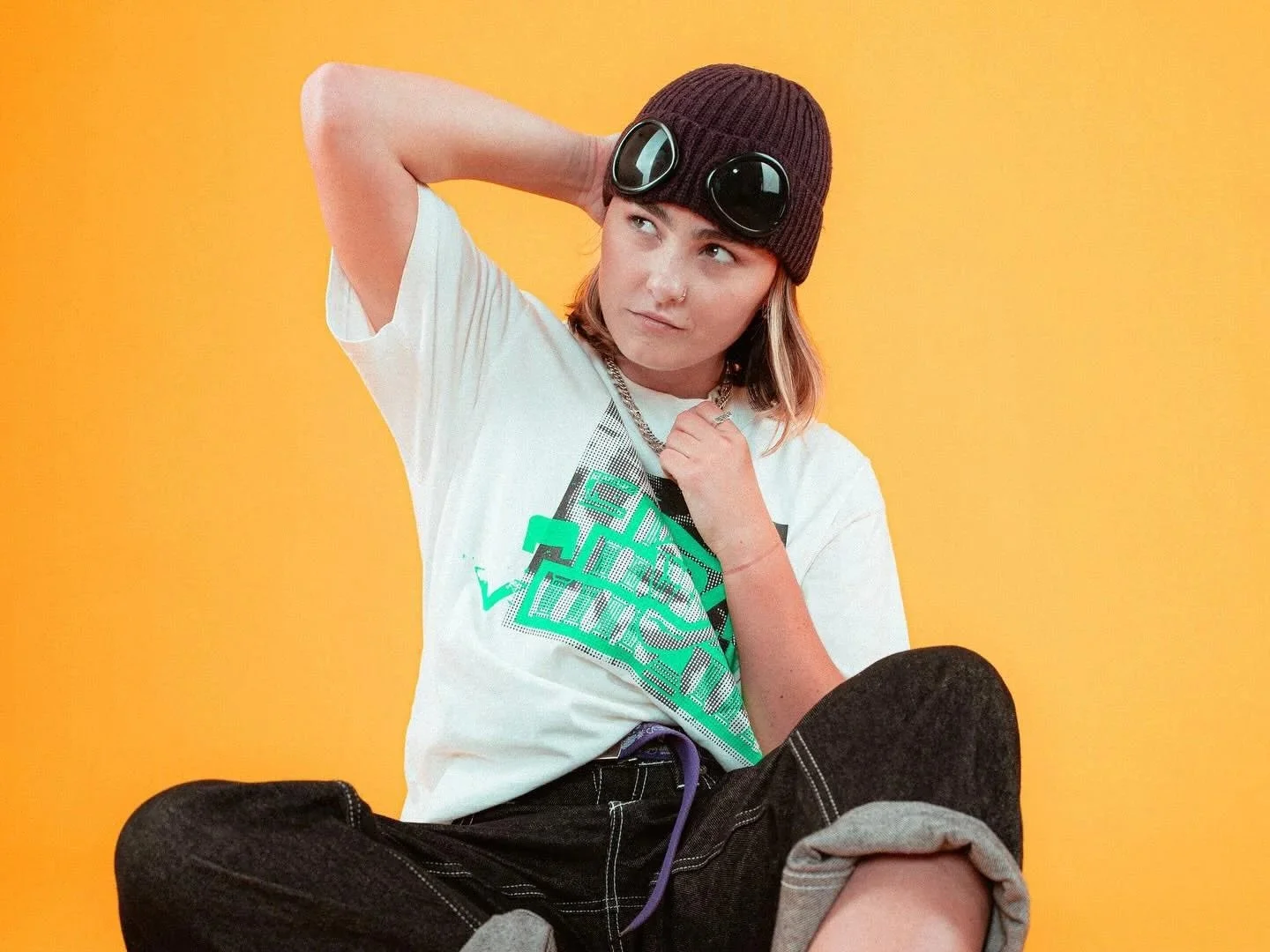Young woman wearing a brown beanie with goggles on it, a white graphic t-shirt, and black jeans, sitting against a yellow background, looking thoughtfully to the side.