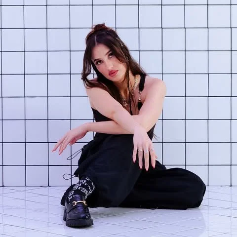 Woman with dark hair in a black outfit sitting on the floor against a tiled wall.