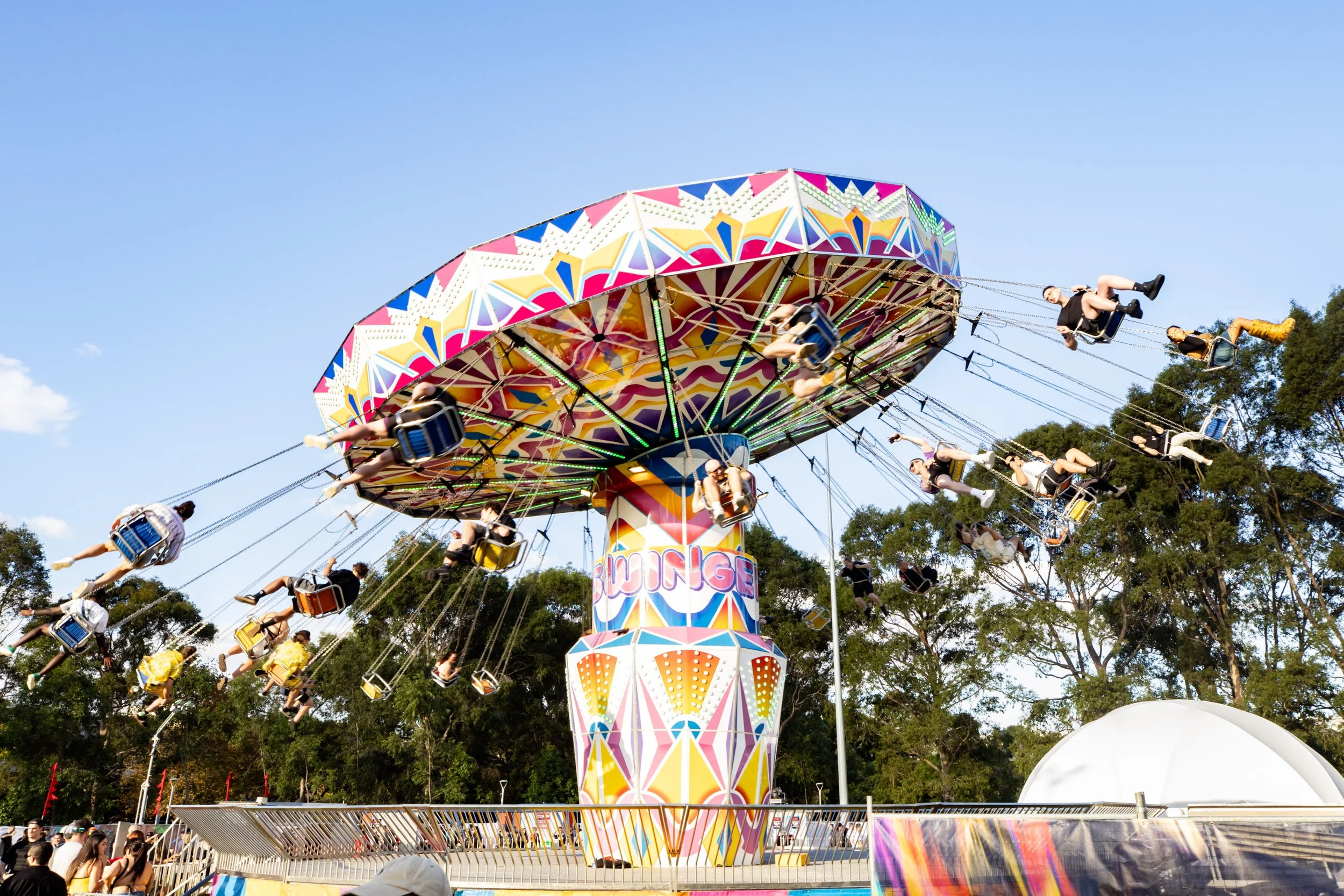 Colorful amusement park swing ride with multiple seats and people riding, spinning in the air, set against a background of trees and a clear blue sky.