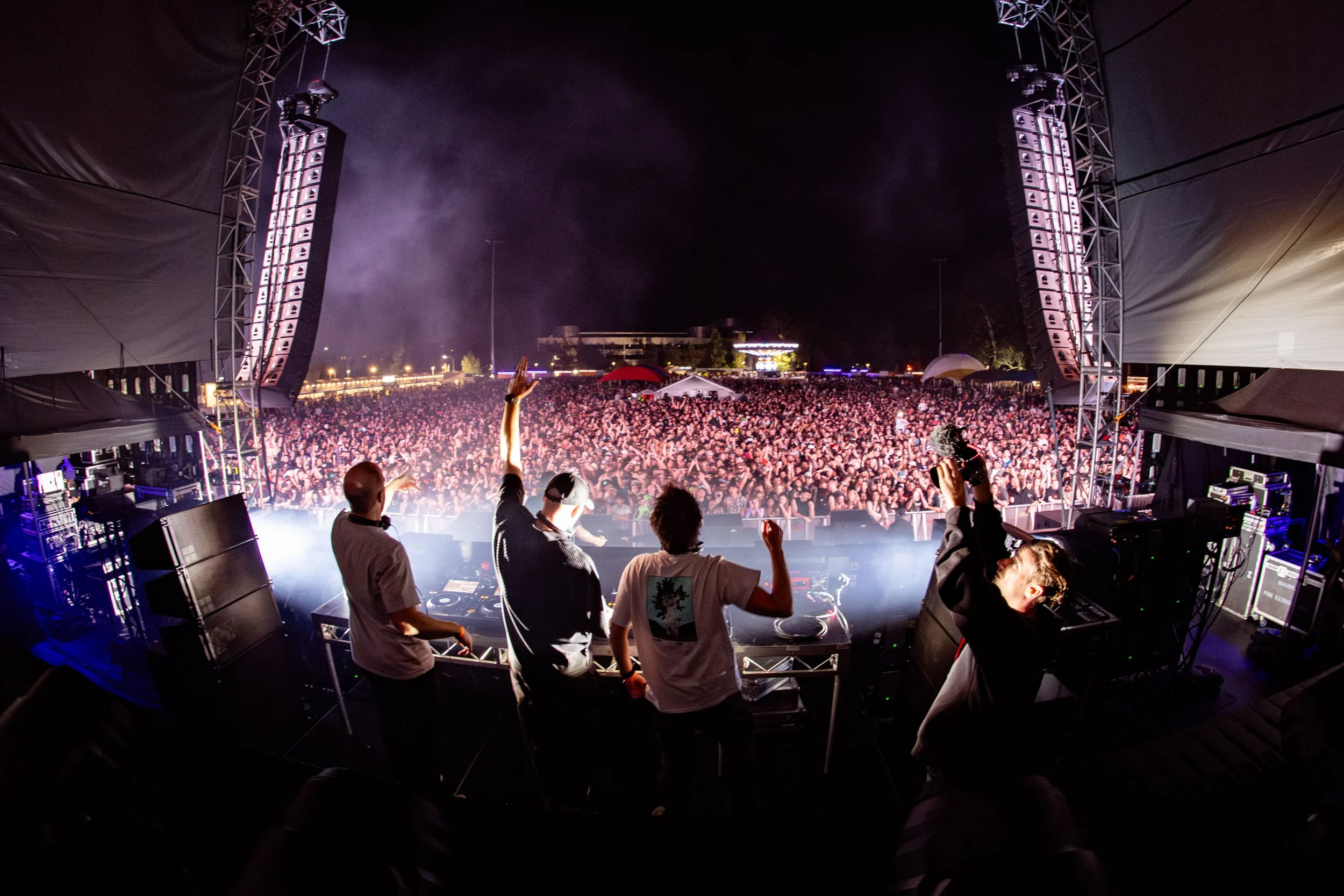 View from a DJ booth showing four DJs performing at an outdoor music festival at night. The crowd extends into the distance, illuminated by stage lights, with smoke and stage equipment visible.