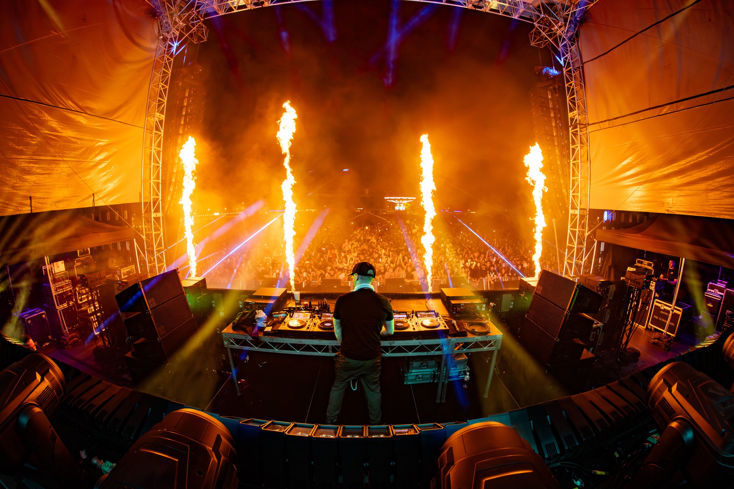 DJ performing on stage with fire pyrotechnics at a music festival. A large crowd is in front of the stage under colorful lights.