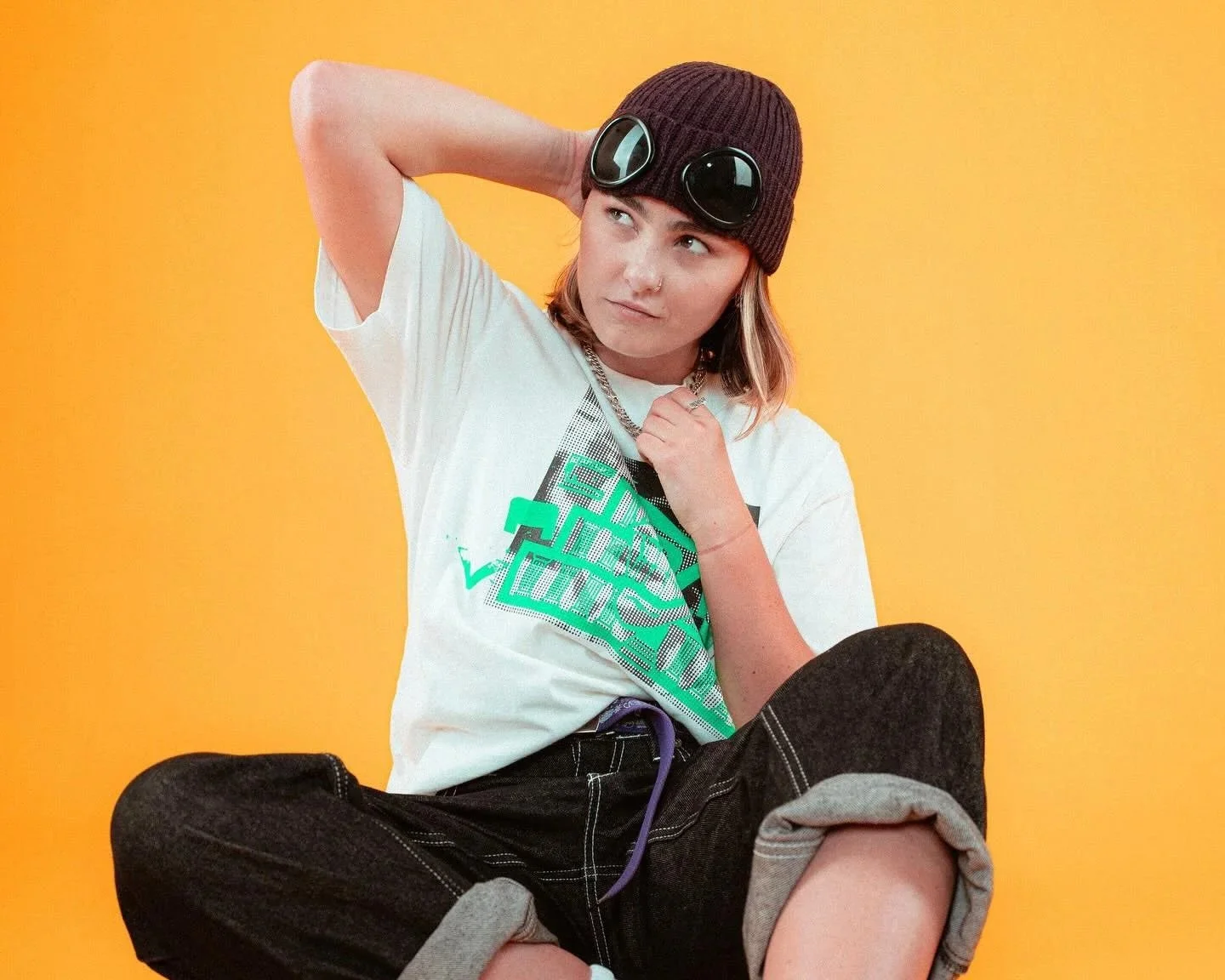 Young woman sitting against yellow background, wearing a dark beanie with glasses, a white graphic t-shirt, and black shorts, posing with one hand behind her head and the other near her neck.