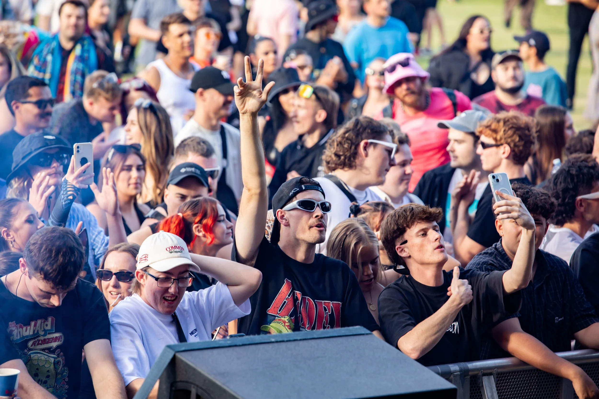 Crowd of people at outdoor event, some taking photos and videos, with a person in the front making a peace sign.