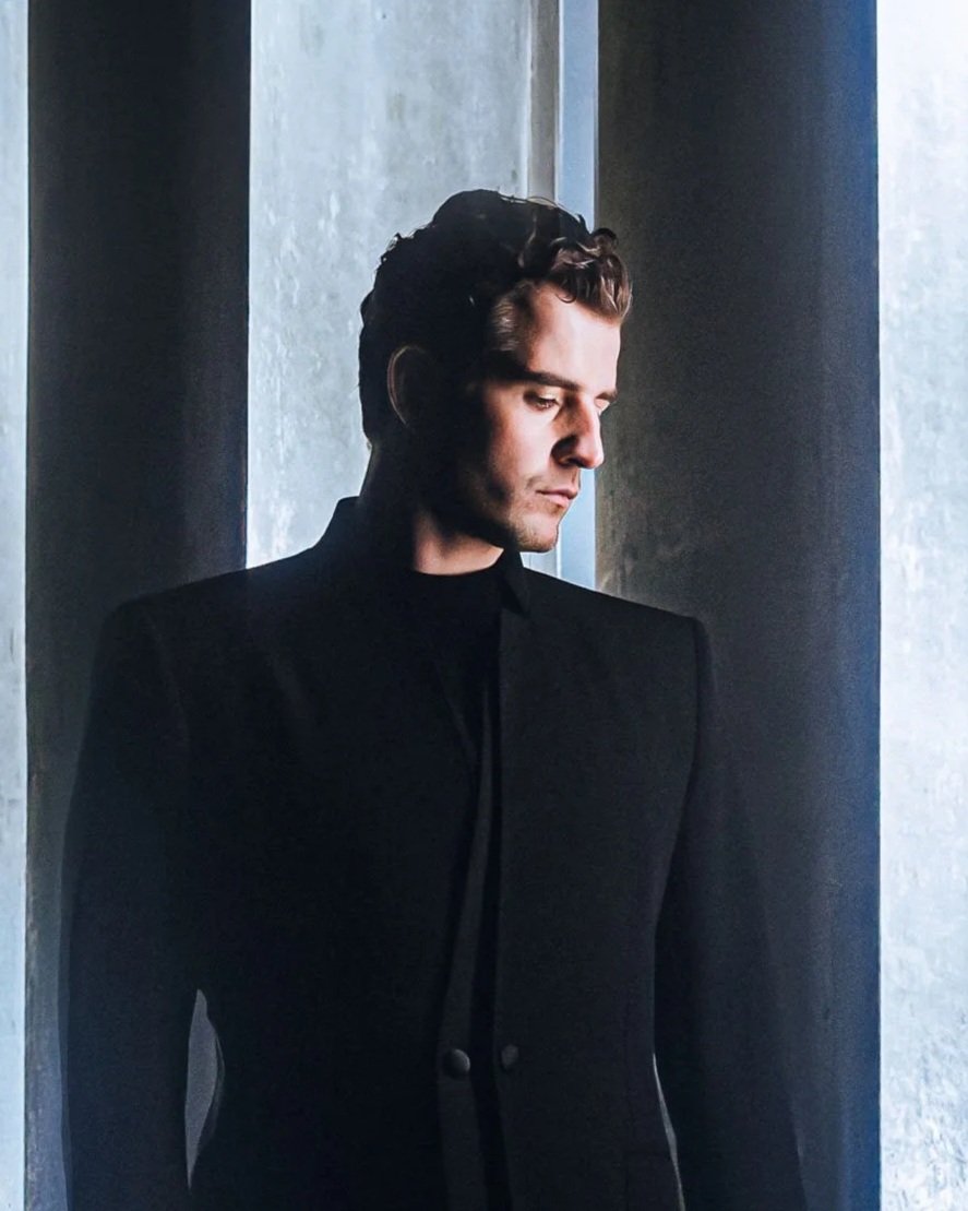 A man with dark, wavy hair and light skin, wearing a black blazer and shirt, standing near a textured wall, looking down with a serious expression.