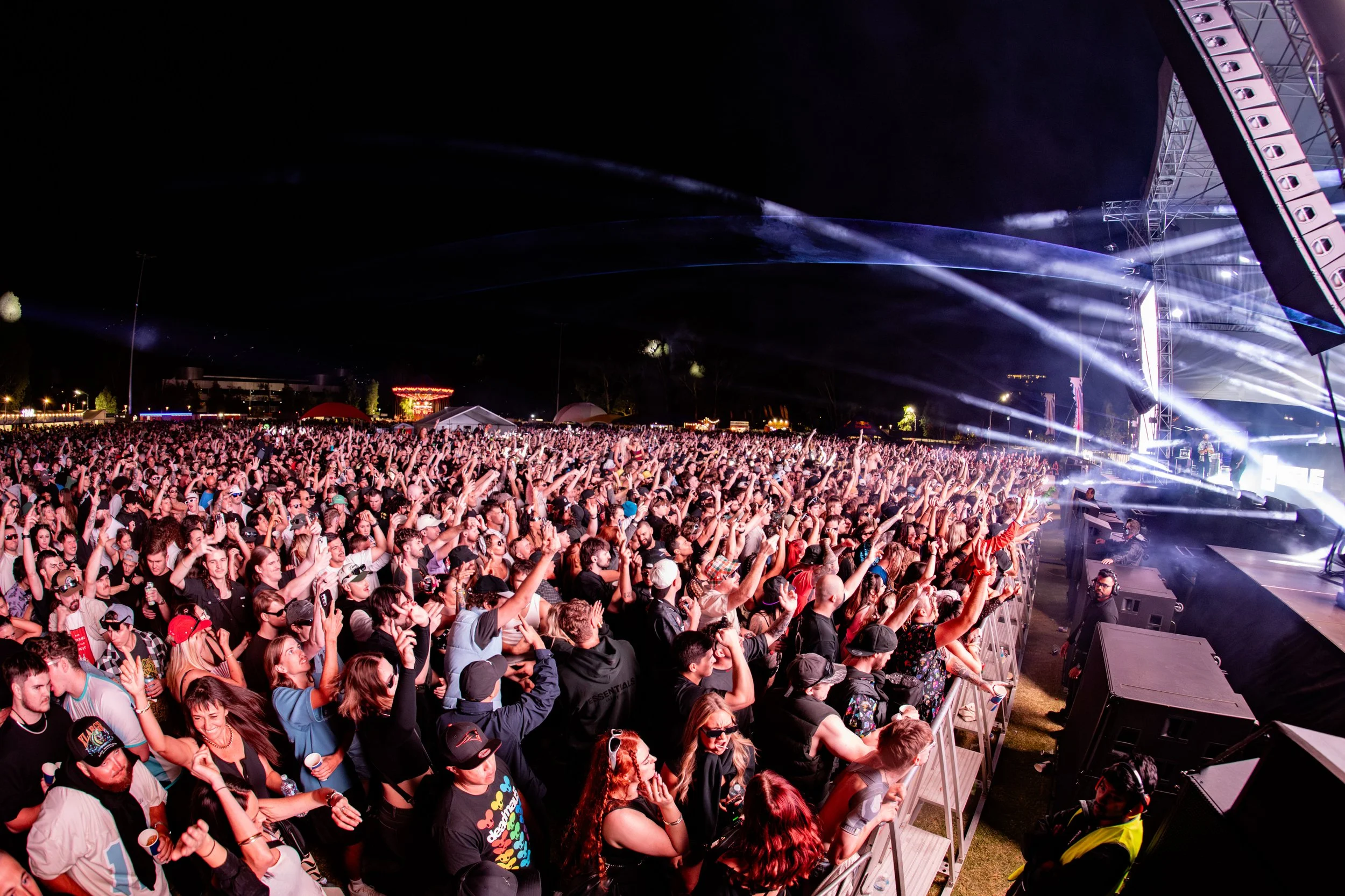 Large crowd of people at an outdoor concert, facing a stage with bright lights and speakers, at night.