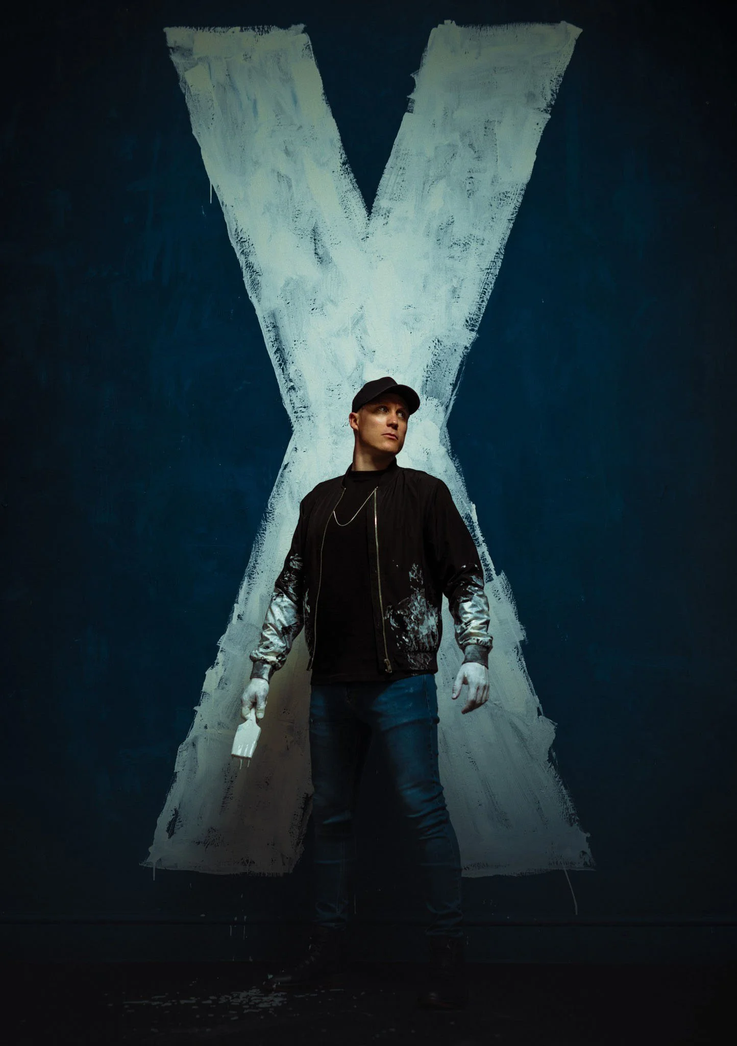 A person in casual clothing stands in front of a large painted X on a wall.