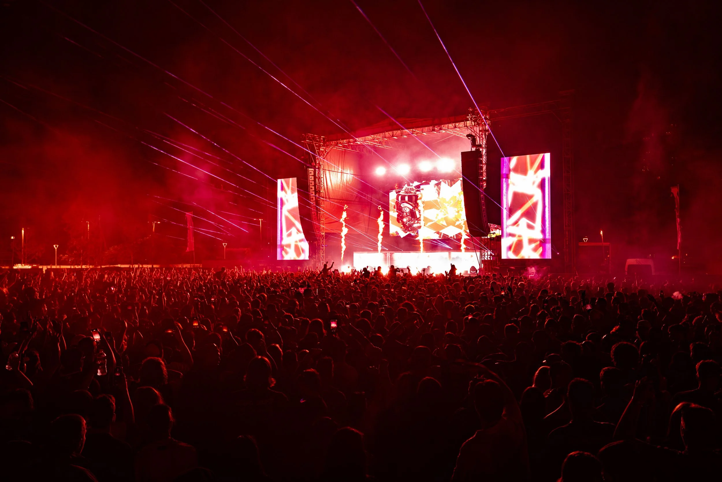 Crowd of people at a concert or music festival during the night, with a bright stage featuring large vertical LED screens and pyrotechnics, all bathed in red lighting.