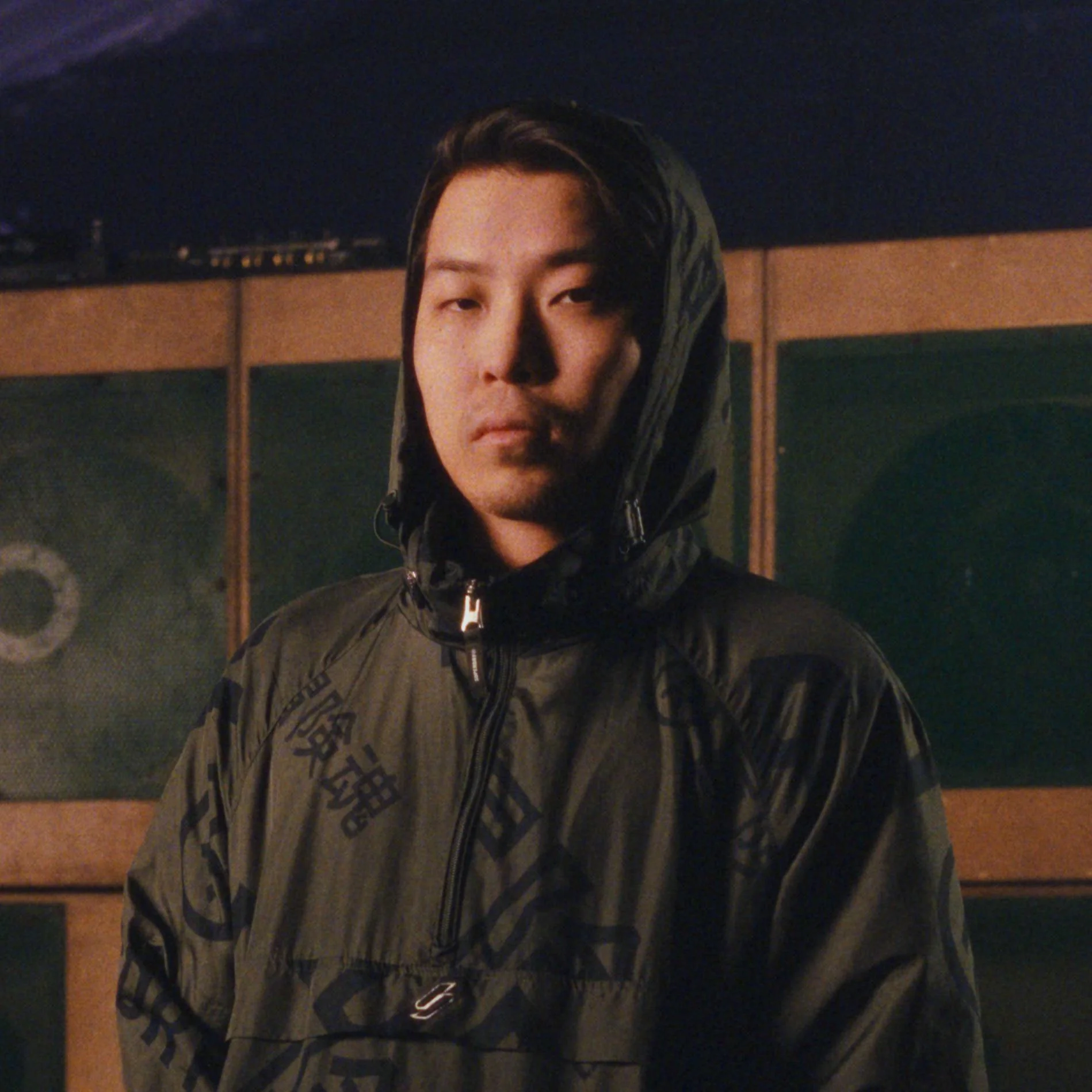 A young man with medium-length dark hair wearing a black hooded jacket, standing in front of a wooden sound system with speakers.
