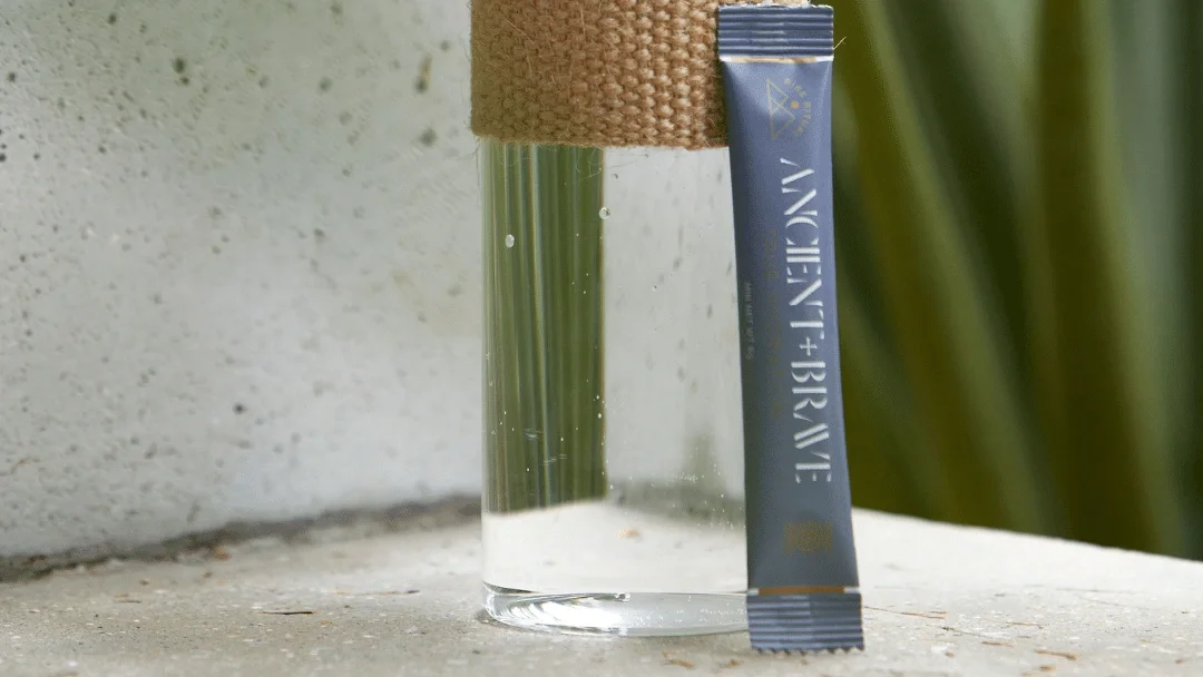 An Ancient + Brave single-serve sachet leaning against a glass water bottle on a pale stone ledge, greenery blurred behind.