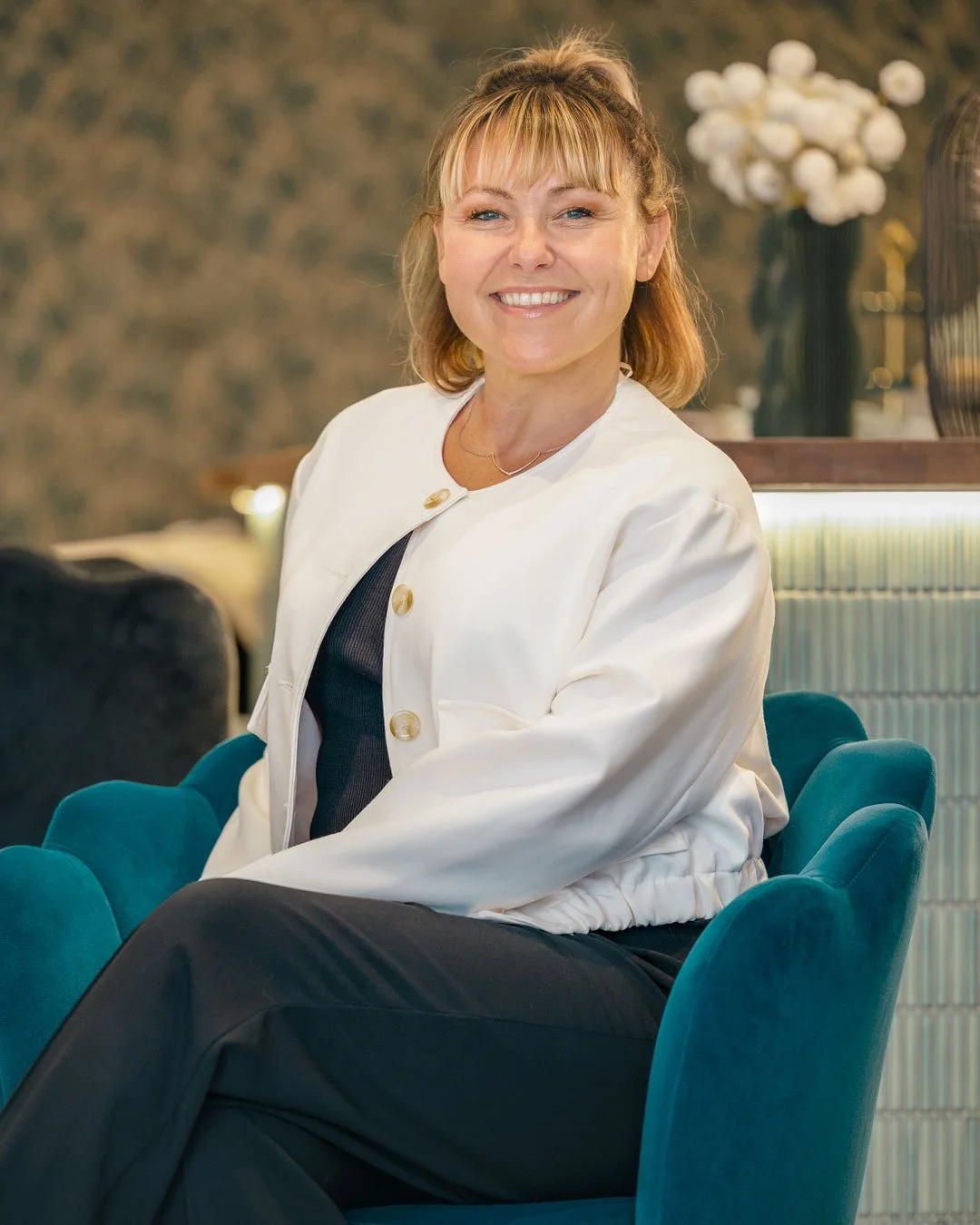 A smiling woman with blonde hair wearing a white jacket and black top, sitting on a teal upholstered chair in a stylish room with a decorative background and a flower arrangement.