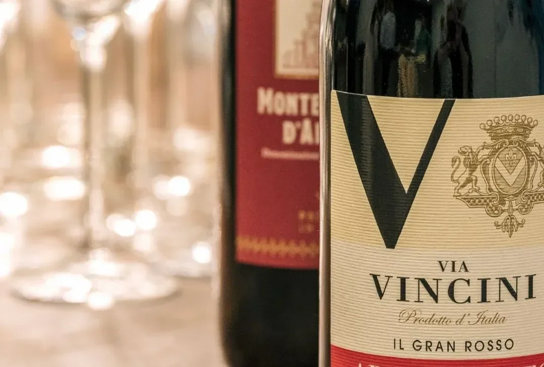 Two bottles of red wine on a wooden table, a row of empty wine glasses softly blurred behind them.