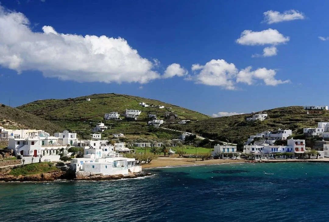 Whitewashed houses around a small sandy bay on Sifnos, with green hills behind and calm blue water in front.