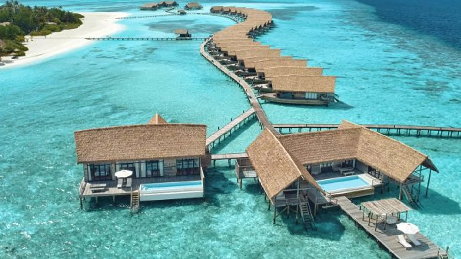 A line of thatched overwater villas on slim wooden stilts above a still turquoise lagoon at soft morning light, a long jetty leading out to sea.