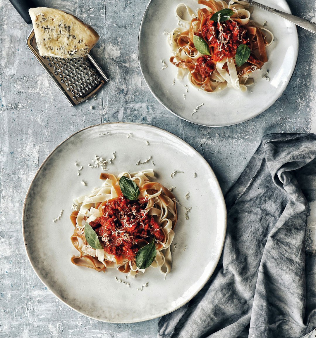 Two plates of fettuccine pasta topped with tomato sauce, grated cheese, and basil leaves, on a textured gray surface with a gray cloth, a wedge of blue cheese, and a metal grater nearby.