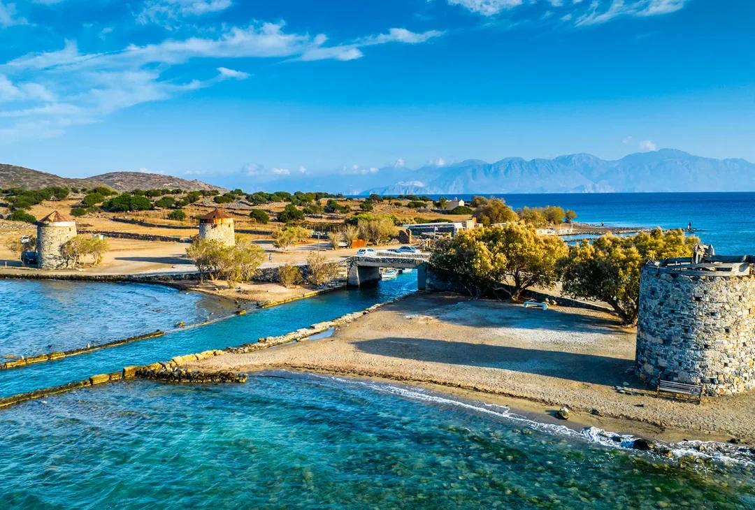 A sunlit Cretan coastline in late autumn, tavernas open along the front, a handful of visitors still out walking.