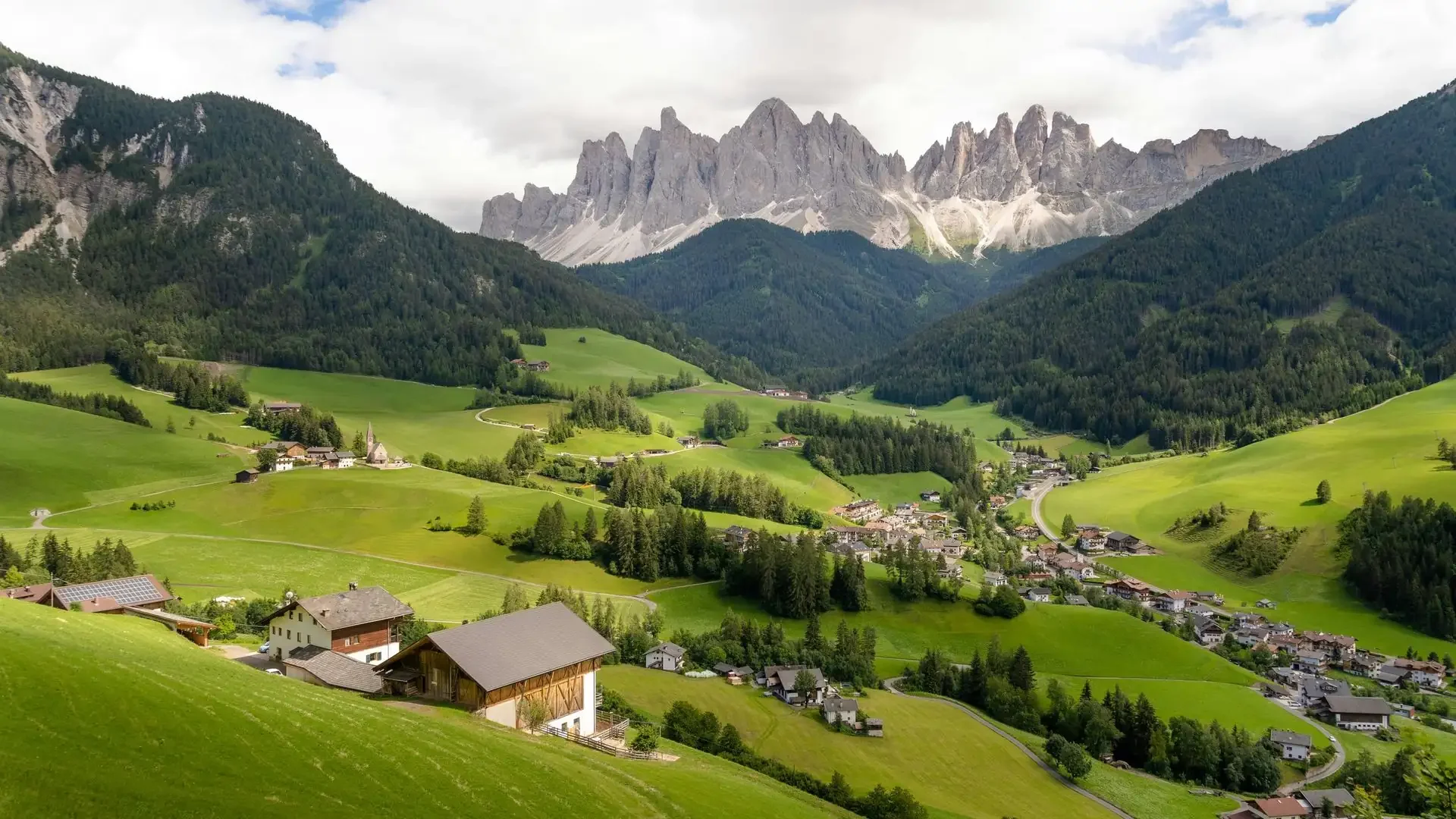 Wide editorial view of Val di Funes valley in South Tyrol, with rolling green meadow, traditional alpine farmhouses, and the jagged Geisler peaks rising behind.