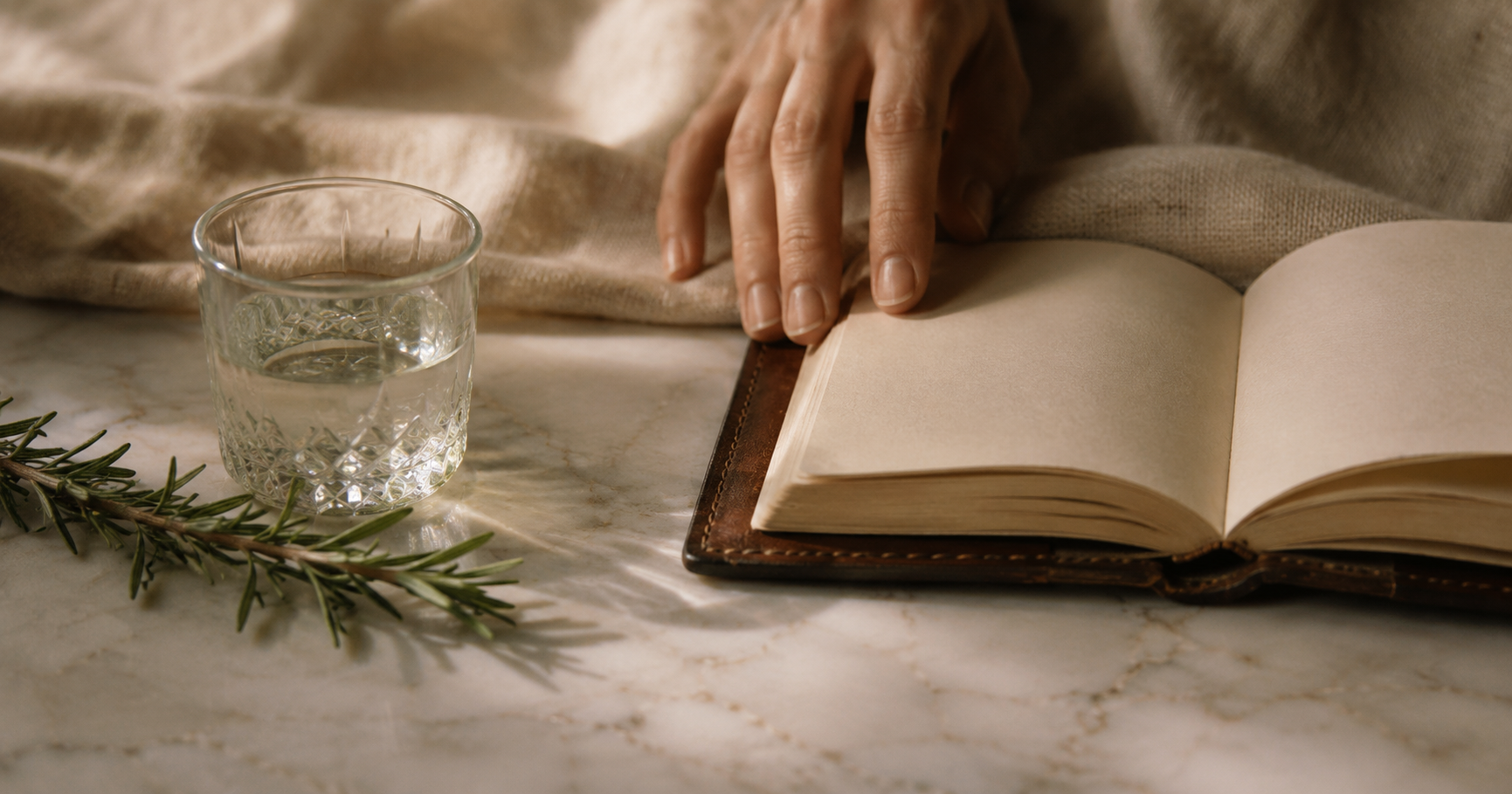 A hand rests on a pale marble surface beside an open leather notebook, a glass of water, and a sprig of rosemary.