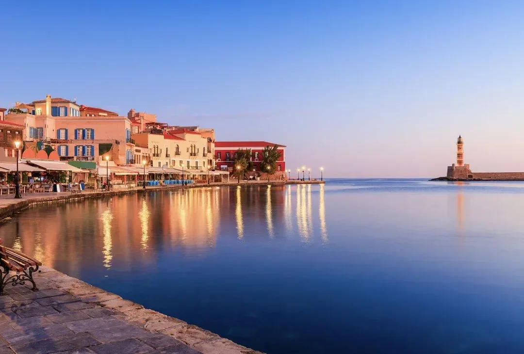Chania's Venetian harbour in west Crete at dusk, its old lighthouse and waterfront lights reflected in the still water.