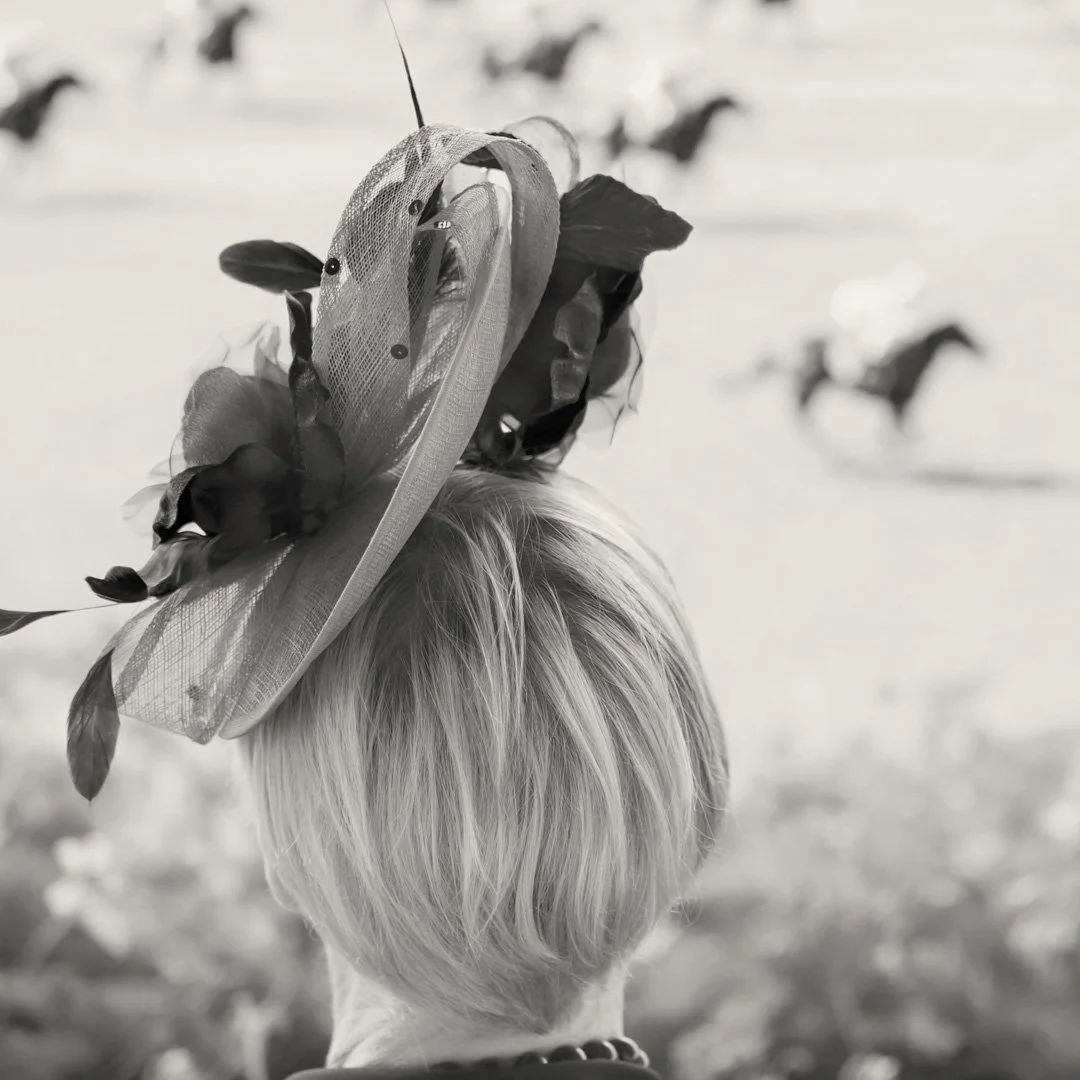 A person with short blonde hair wearing a large decorative hat with flowers, viewed from behind at a horse race, with horses running in the background.