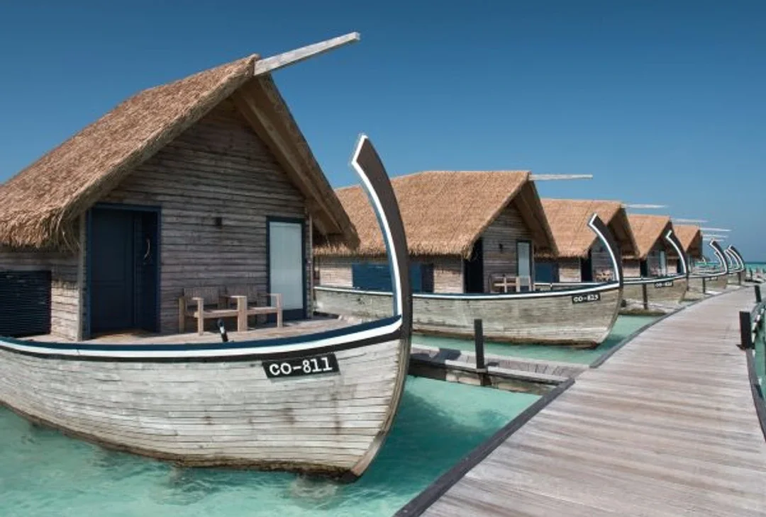 The interior of an overwater suite with a curved boat-shaped ceiling in pale timber, an open deck beyond framing the lagoon.