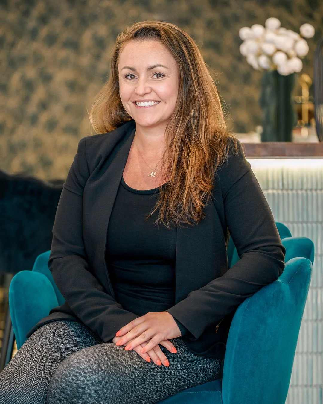 A woman with long, wavy brown hair smiling and sitting on a teal chair in an upscale interior with a floral arrangement in the background.