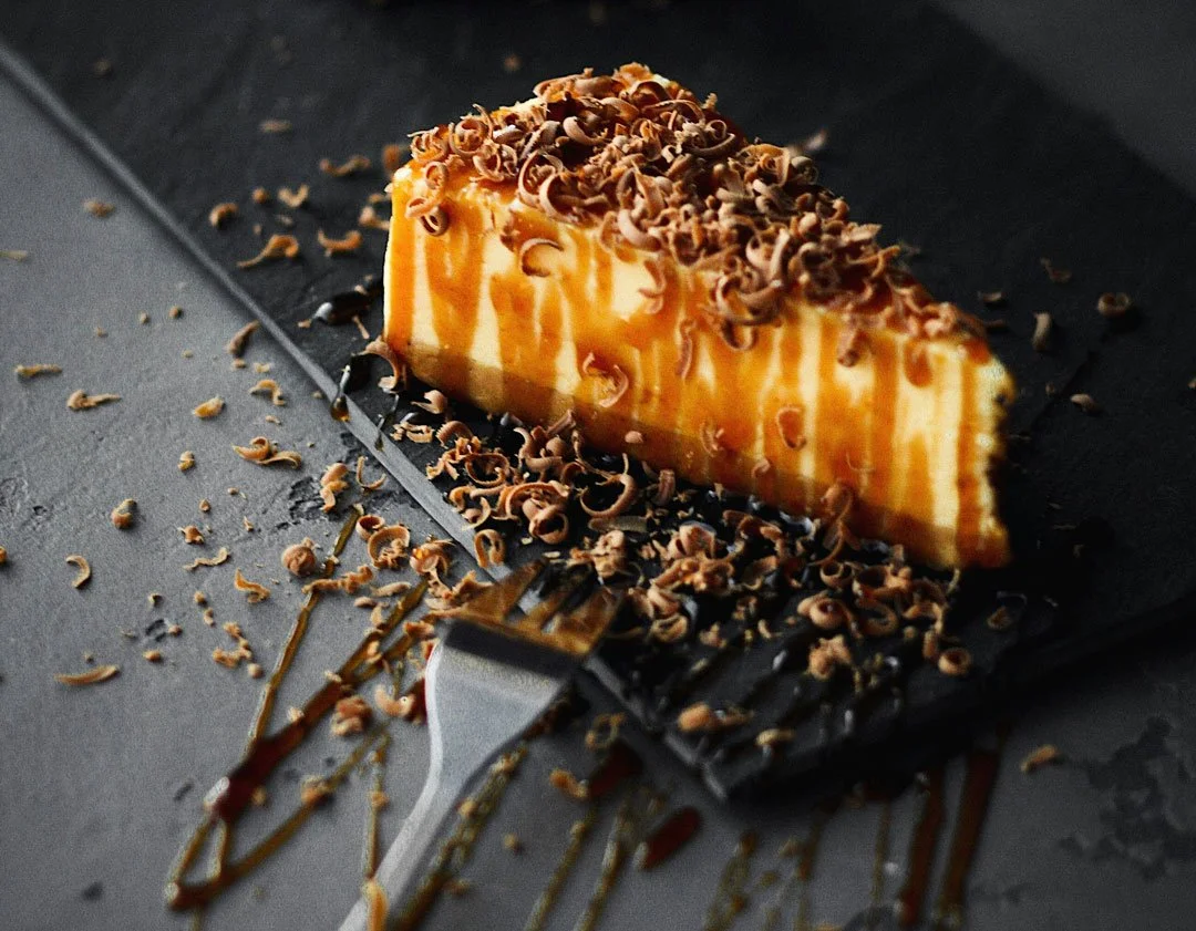 A slice of cheesecake topped with chocolate shavings and caramel drizzle, served on a black slate plate with some chocolate shavings and caramel drizzles around it.