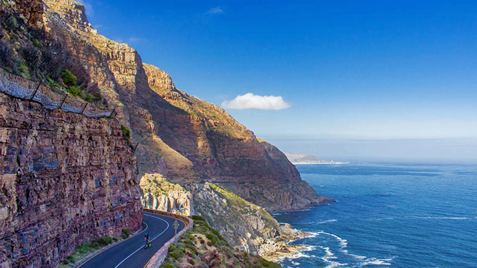 A coastal road curving between dense green forest and a still lagoon, blue mountains rising behind in soft afternoon light.